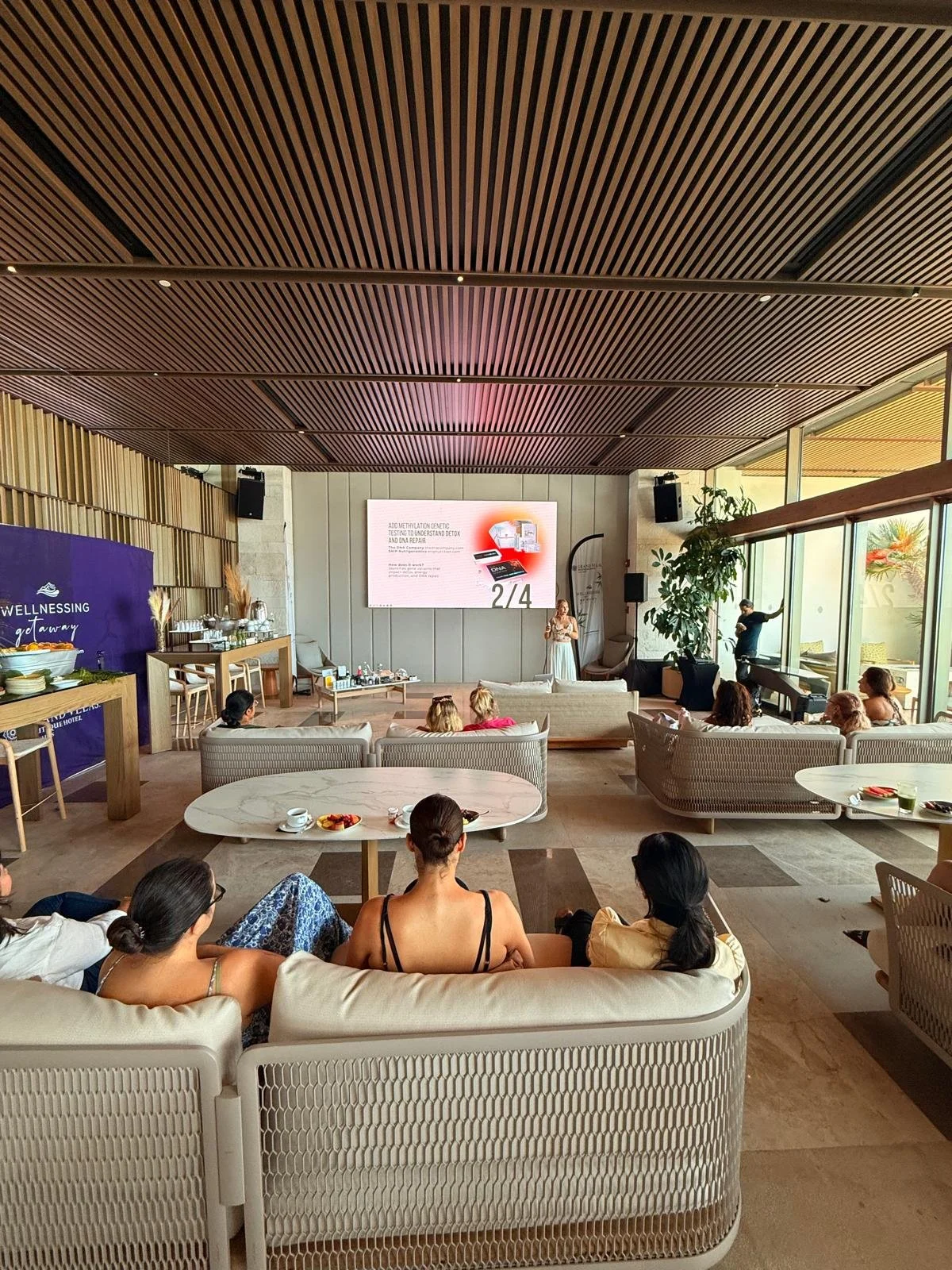A group of people seated on sofas and chairs in a modern lounge or conference room, watching a presentation on a large screen.