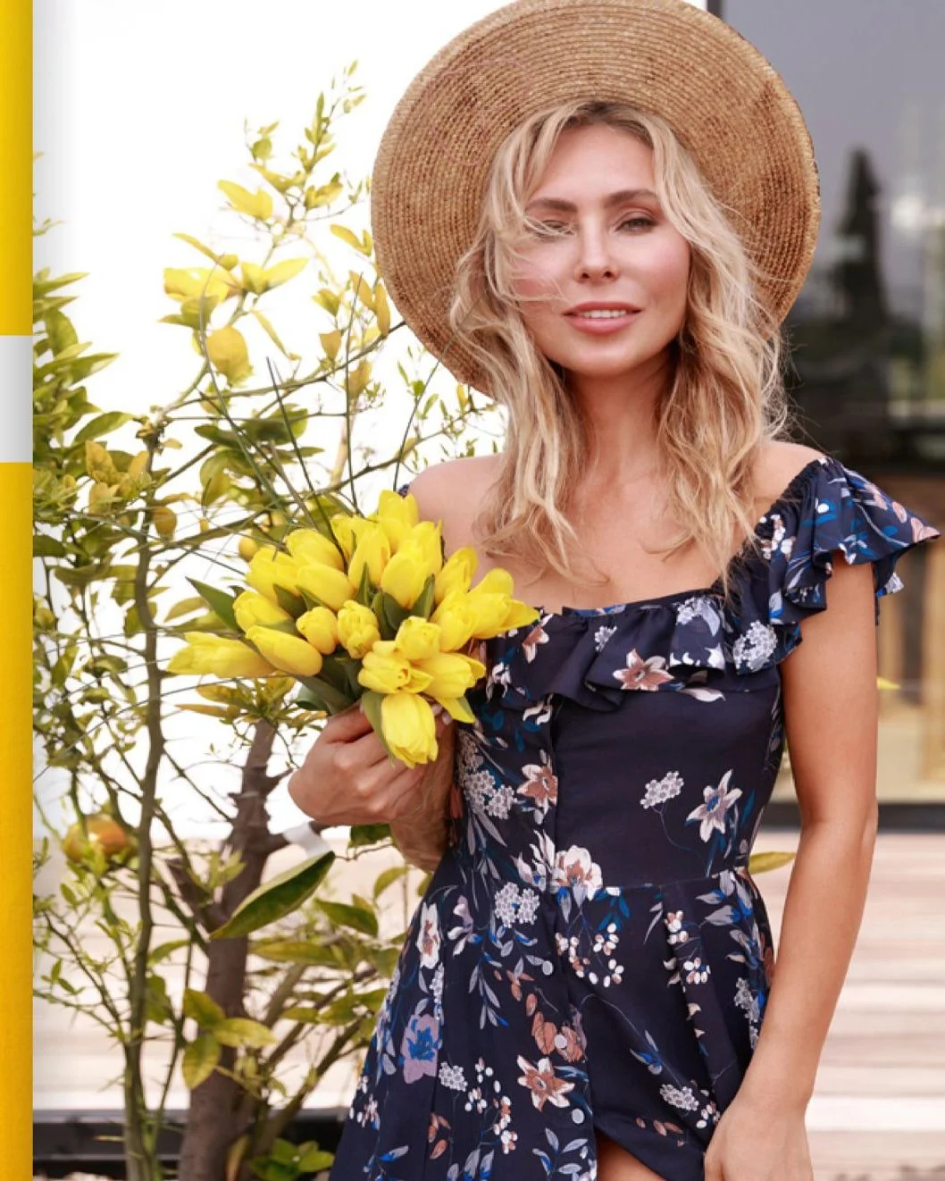 A woman with blonde curly hair wearing a straw hat and a navy blue floral dress holding a bouquet of yellow tulips, standing outdoors with a tree and a building in the background.