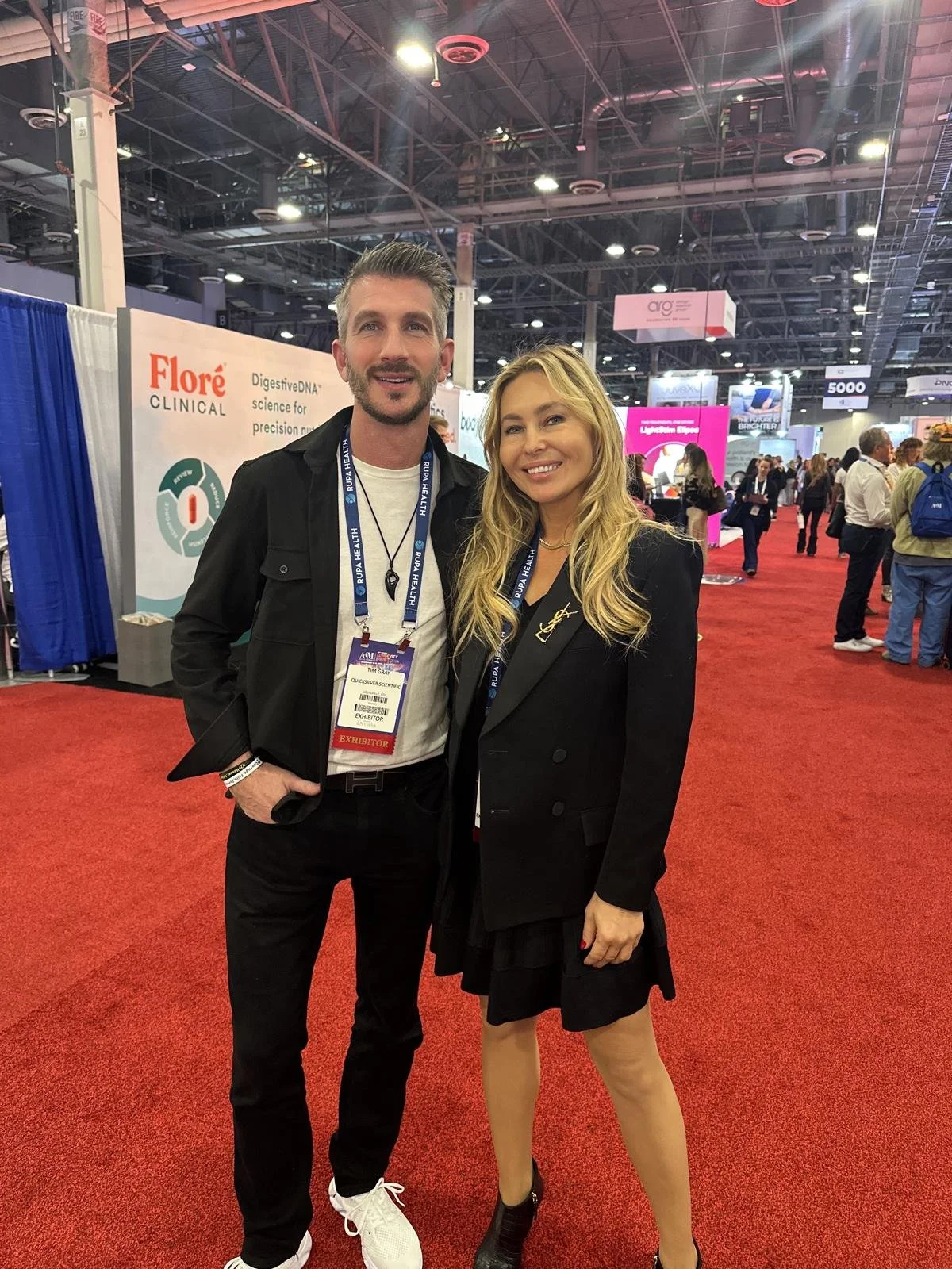 A man and a woman are standing together at a conference, smiling at the camera, with exhibition booths and other attendees in the background.