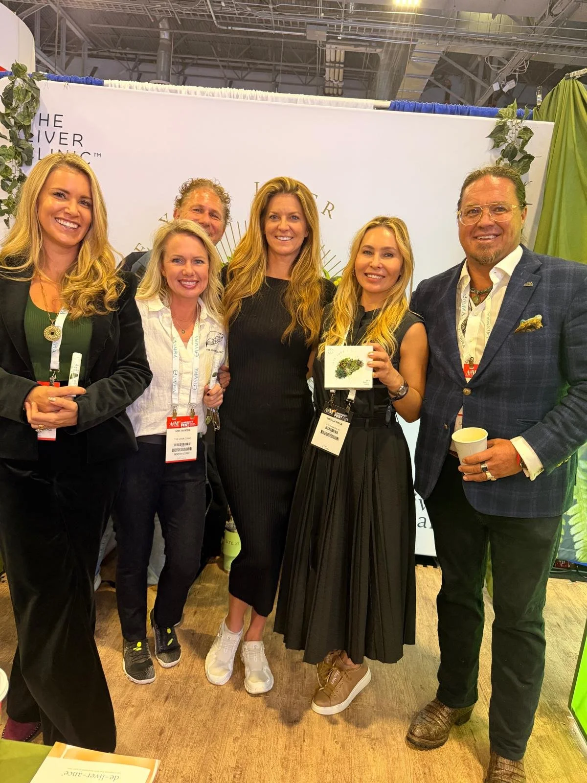 A group of six smiling adults standing together at an event, with some holding cups and a small box, in front of a white backdrop with writing and greenery.