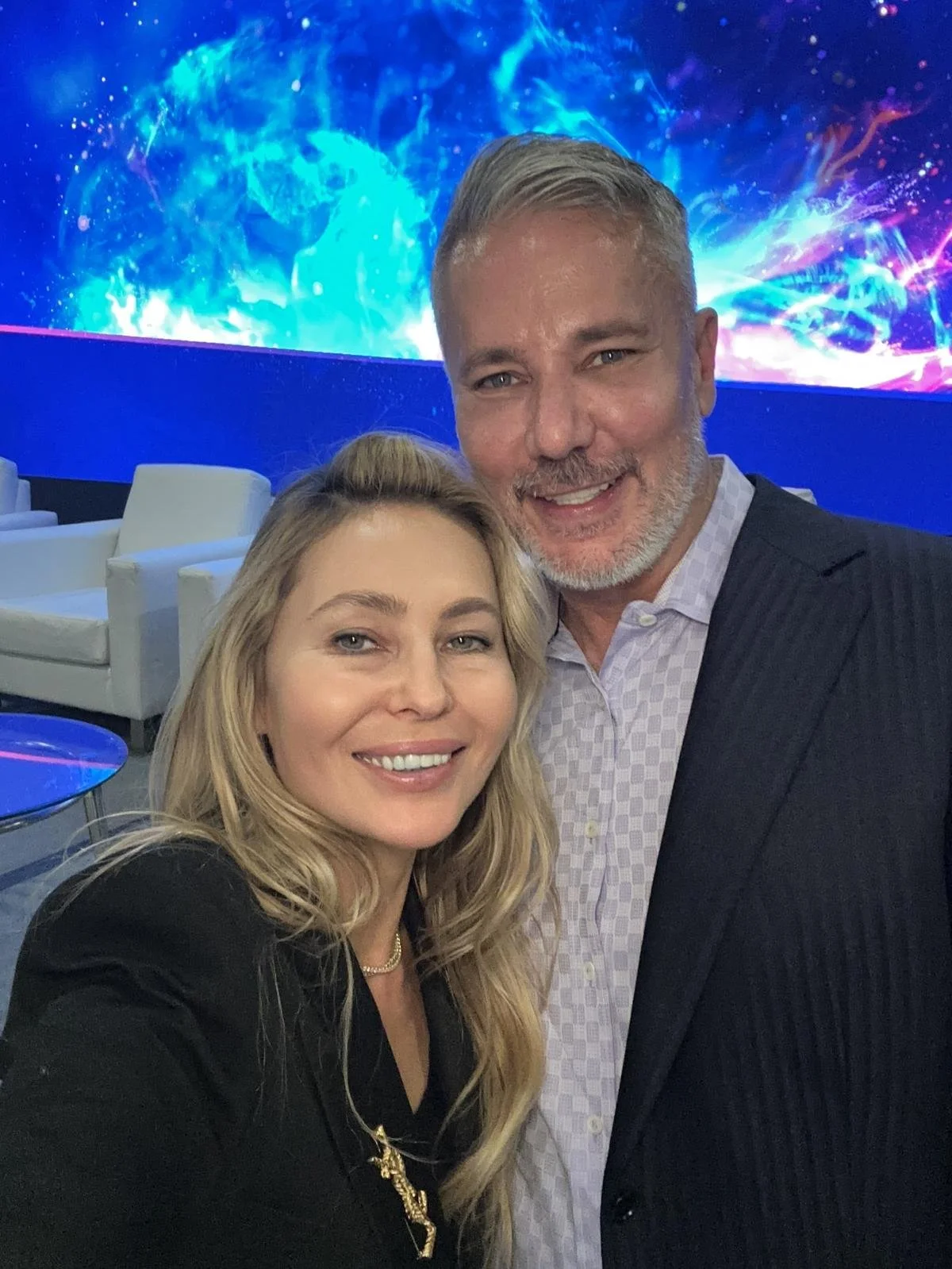 A man and woman taking a selfie together at an event with a digital blue and purple abstract background.