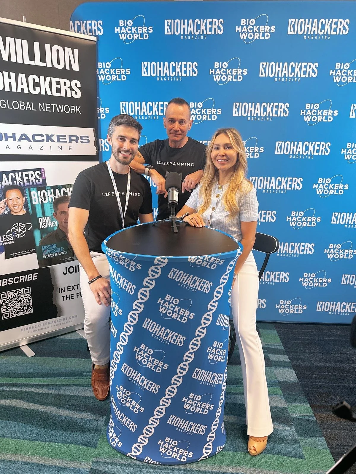 Three people at a BioHackers World event standing behind a table with a microphone, with a blue backdrop and banners promoting BioHackers Magazine. Two men are wearing black shirts with white text, and a woman is wearing a white top and beige pants.