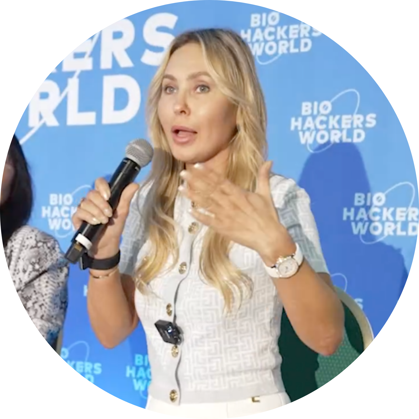 A woman with long blonde hair speaking into a microphone at a conference with a blue backdrop that reads 'Bio Hackers World.'