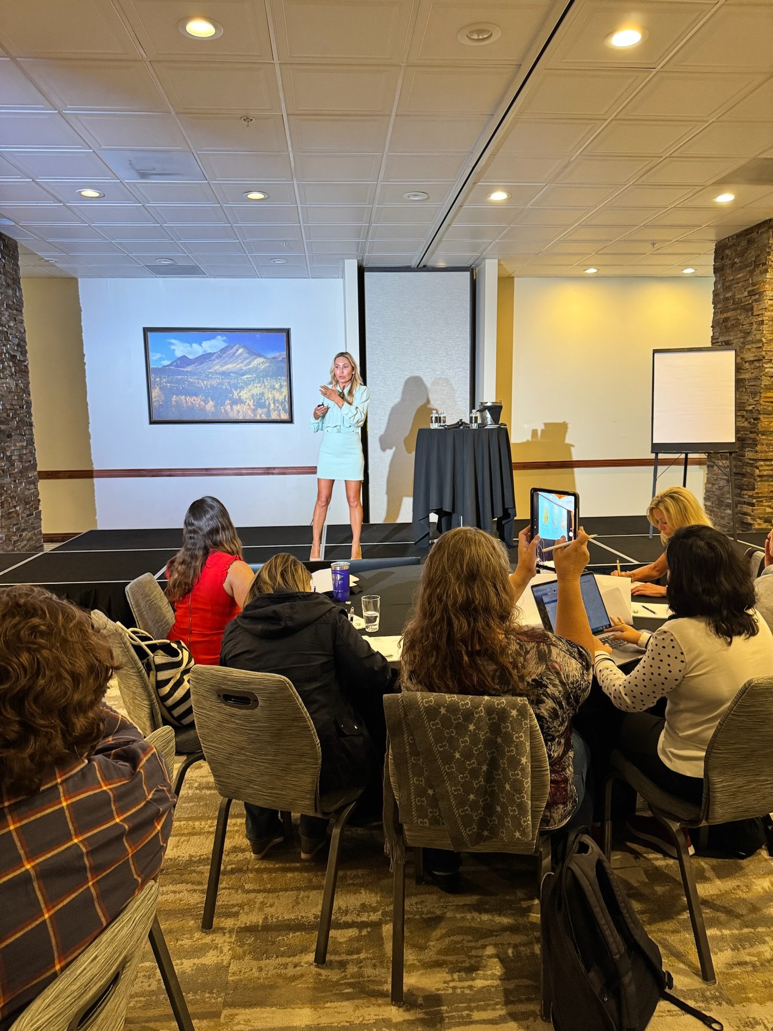 A woman giving a presentation on a stage to an audience in a conference room.