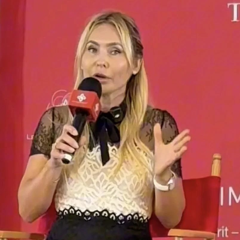 A woman with long blonde hair speaking into a microphone during an interview or panel, seated in front of a red background.