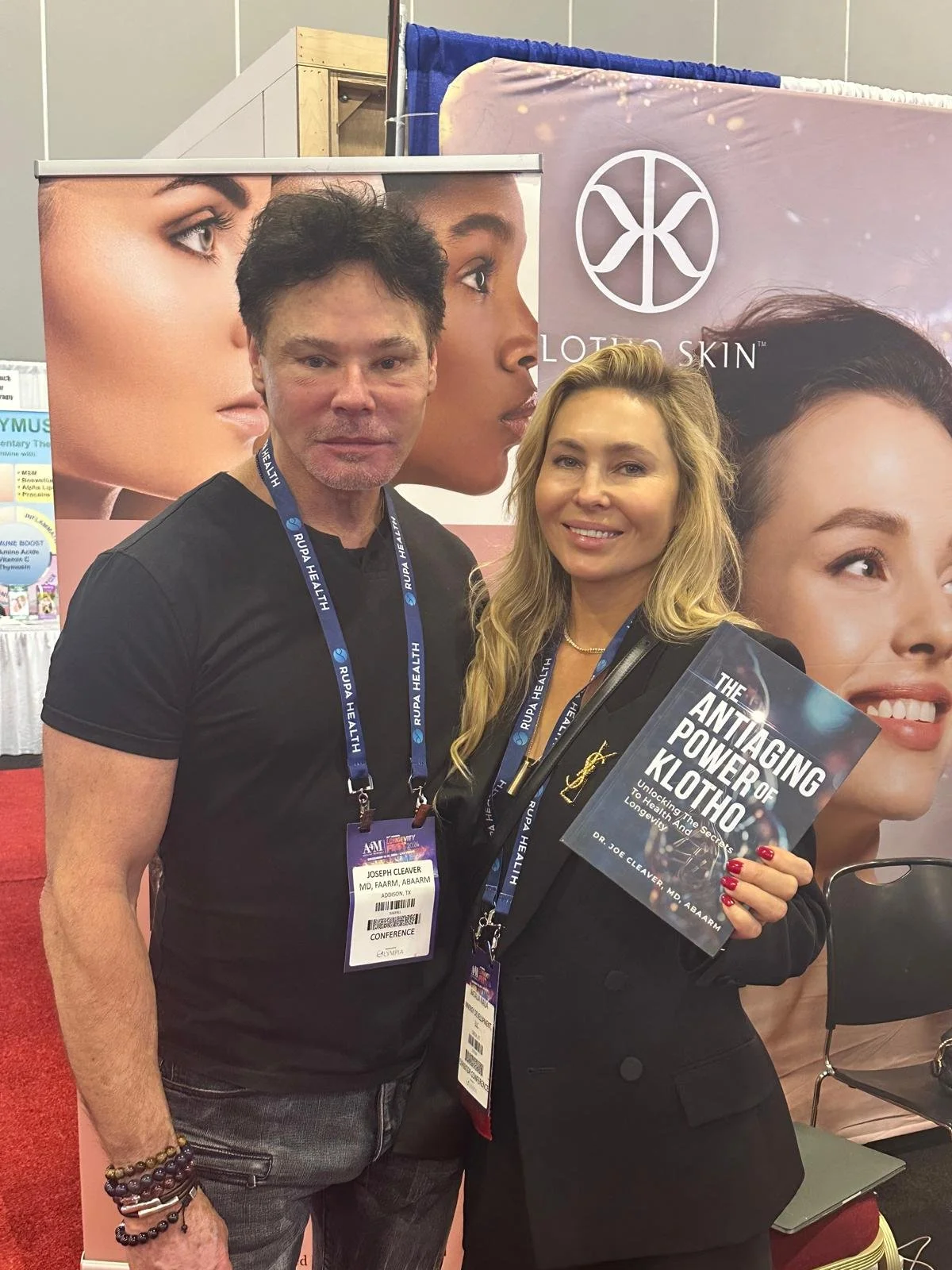 A man and woman are posing together at a conference, holding a book titled 'The Antagonizing Power of Klotho: Unlocking the Secrets to Healthy Aging and Longevity.' They are standing in front of a promotional backdrop with images of diverse women and