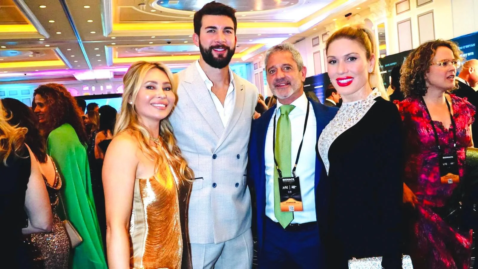 Four people, two women and two men, dressed in formal attire, smiling at a conference or event in a brightly lit, decorated ballroom.