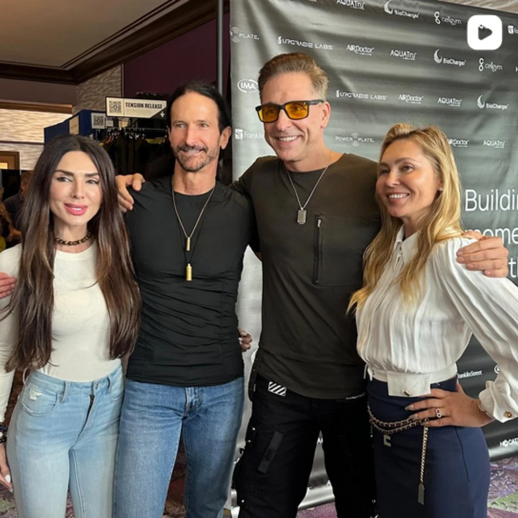 Four people standing together and smiling for a photo in front of a promotional backdrop. The group includes two women and two men, with the man in the middle wearing sunglasses. They are dressed casually, and the setting appears to be at an indoor e