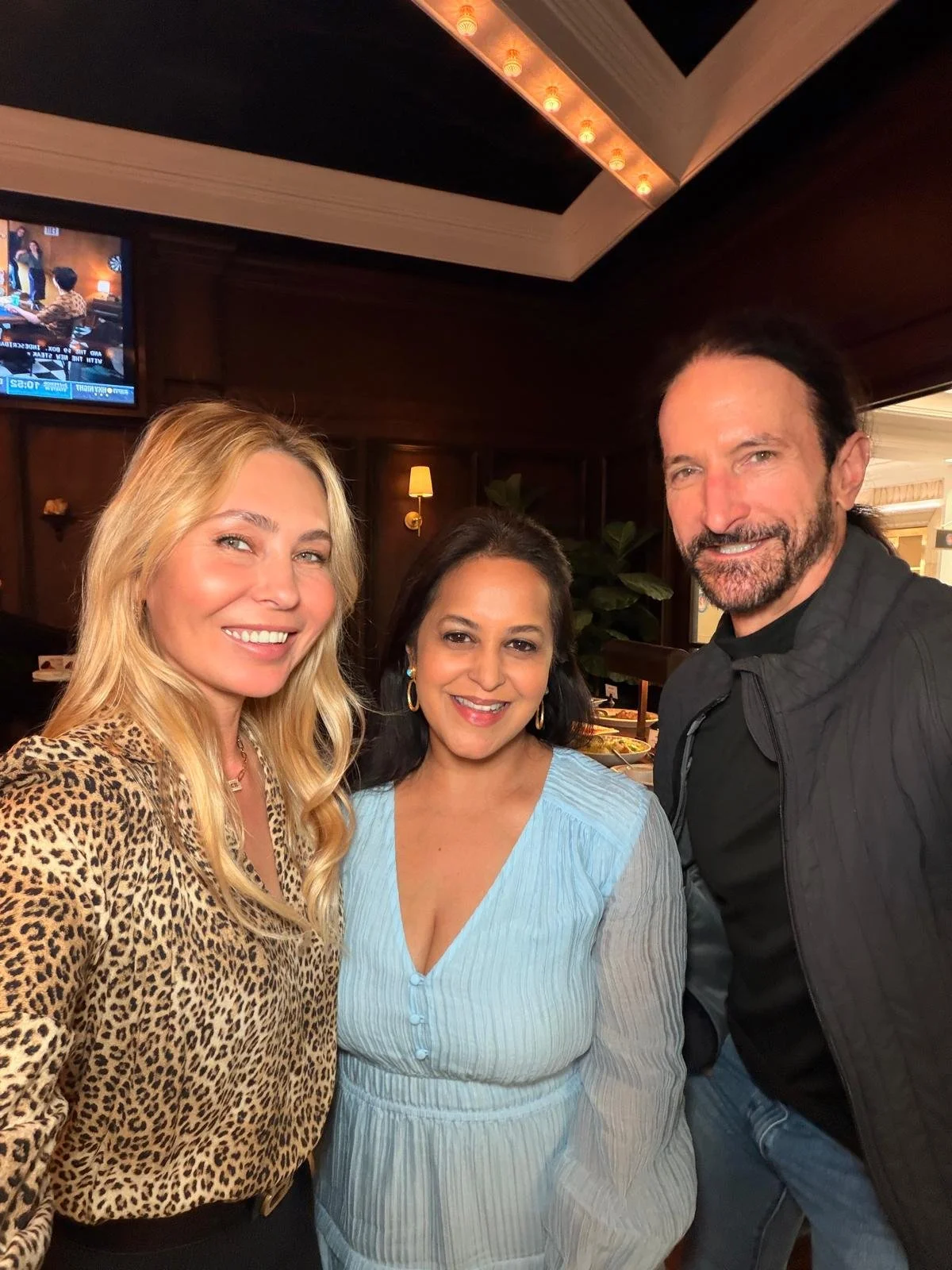 Three people smiling for a photo indoors, with a woman on the left wearing a leopard print top, a woman in the center wearing a light blue blouse, and a man on the right wearing a black shirt and jacket. The background features wood-paneled walls, li