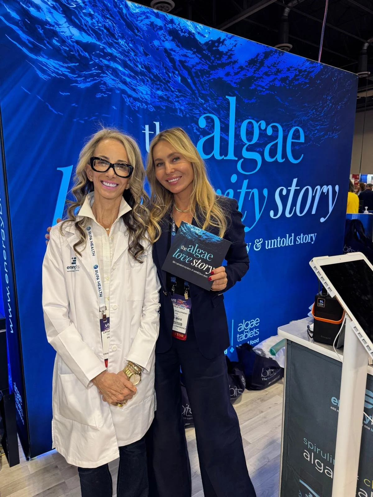 Two women at an event, standing in front of a blue backdrop with the text 'the algae love story.' One woman wears a white coat and glasses, while the other wears a black blazer and holds a booklet. Both are smiling.