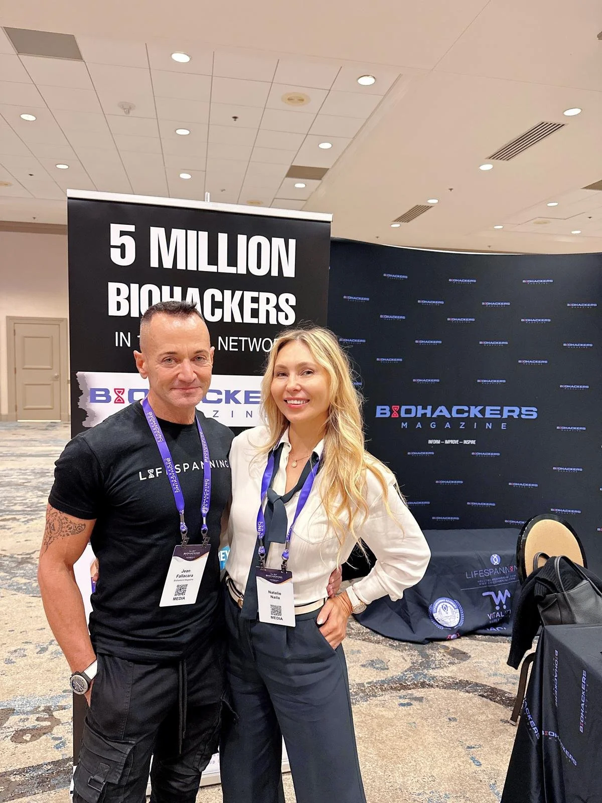 Two people posing in front of a sign that says '5 Million Biohackers in the Network' at a conference, with banners from Biohackers Magazine and LifeSpann. The man is wearing a black T-shirt and the woman is in a white blazer and navy pants.