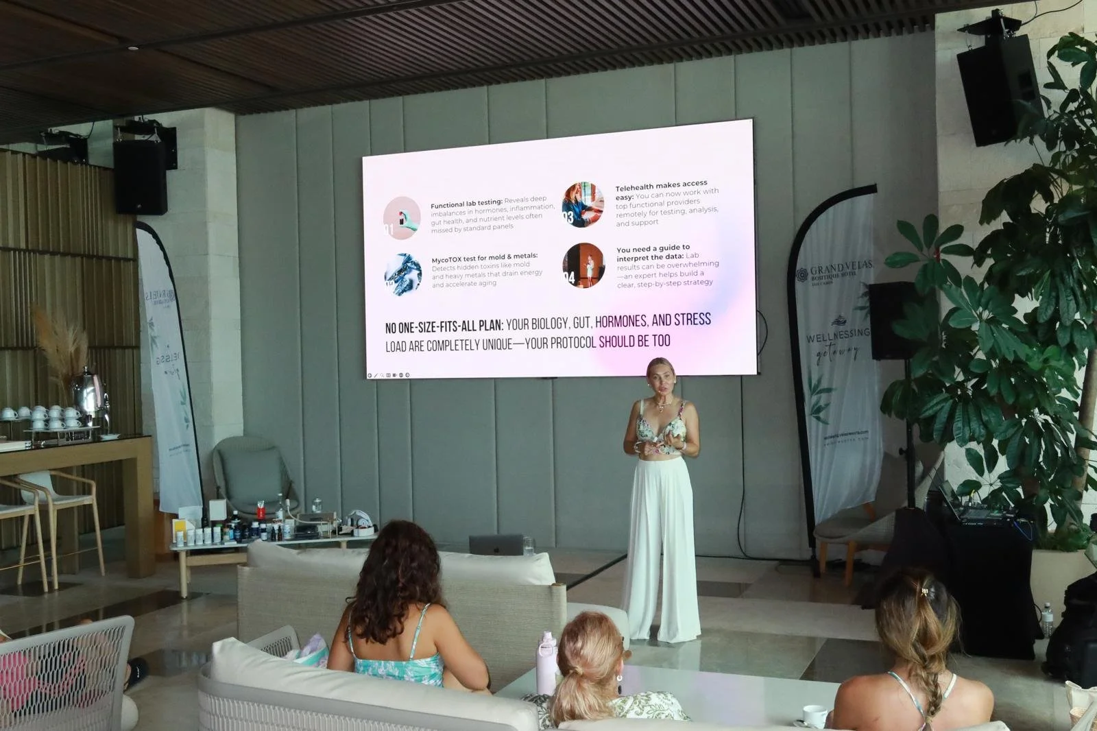 A woman standing and giving a presentation in front of a large screen at an indoor event, with a few women seated in front of her.