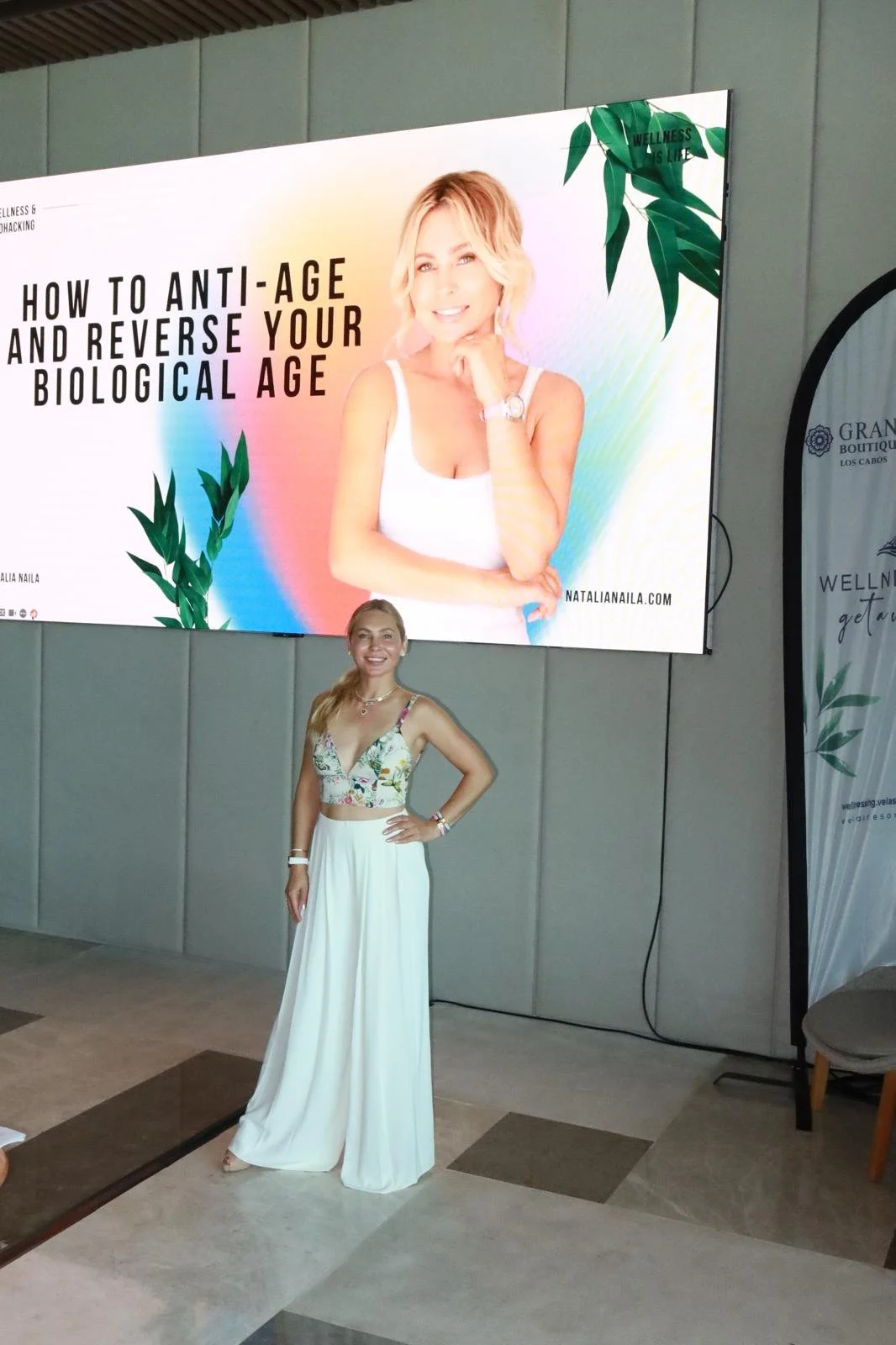 A woman in a white and floral crop top and wide-leg white pants standing in front of a large billboard. The billboard displays a smiling woman with blonde hair and the text 'How to Anti-Age and Reverse Your Biological Age'.