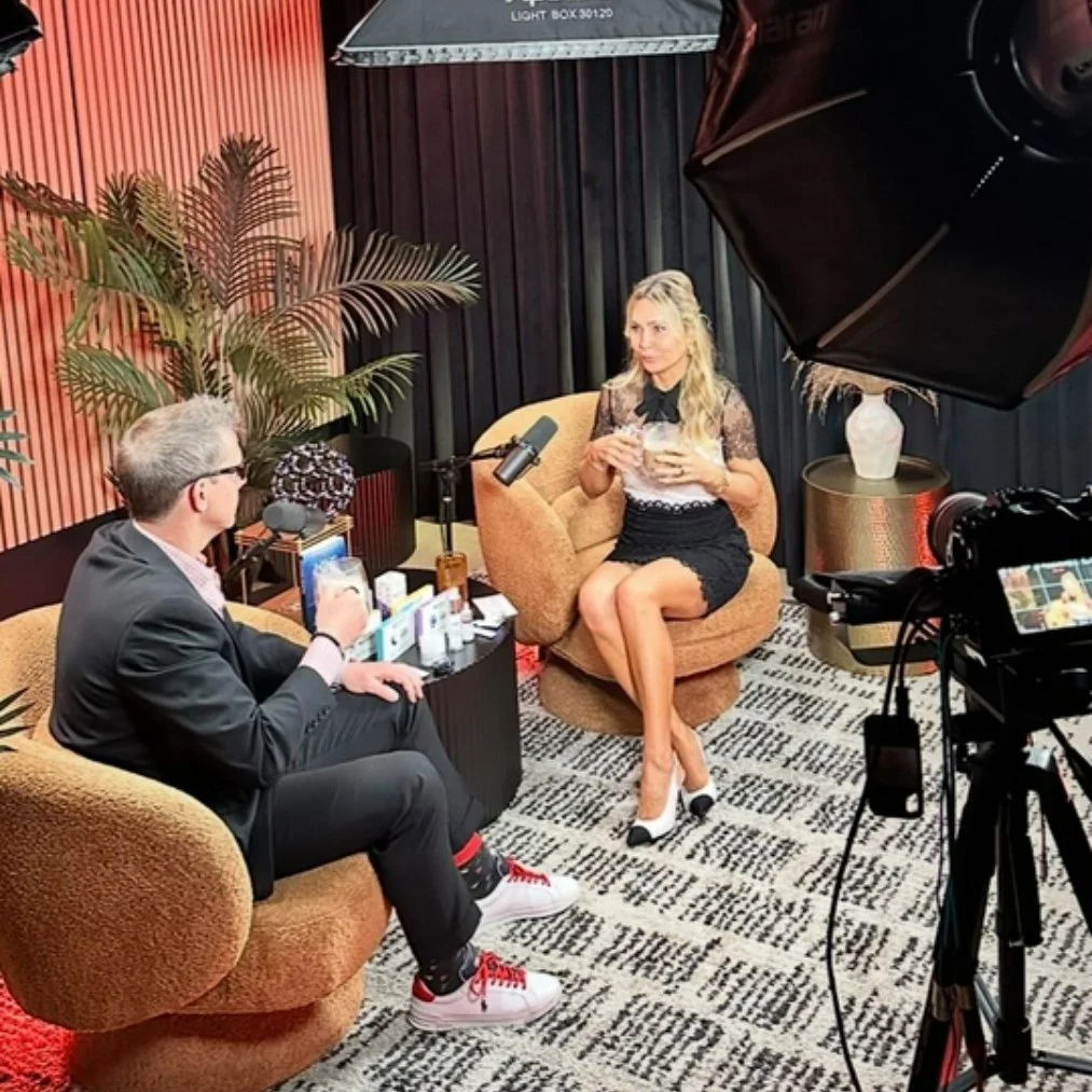 A woman in a black dress with lace sits on a brown chair, holding a drink, with a camera and recording equipment around her. She is being interviewed by a man in a suit with glasses, sitting on a matching brown chair, in a cozy, modern indoor setting