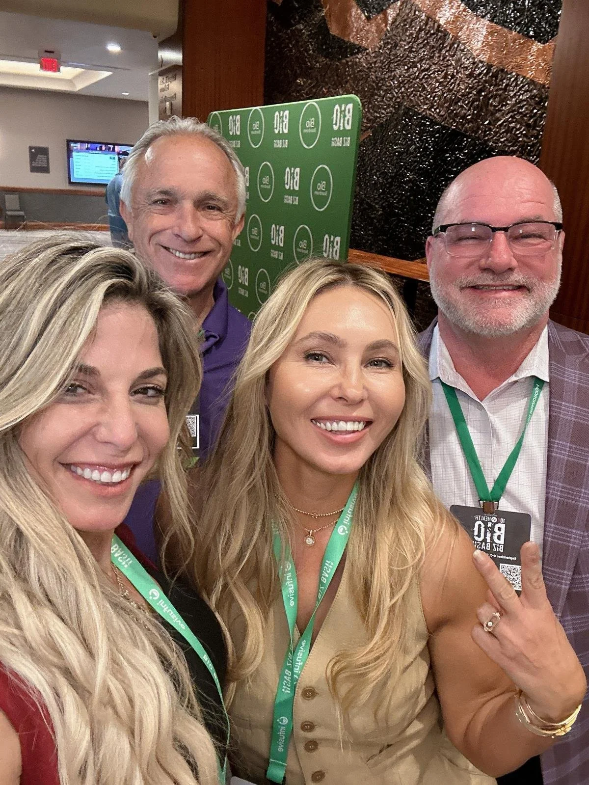 Group of four smiling people taking a selfie at a conference, wearing badges with green lanyards. There is a green backdrop with white logos reading 'BIO.'