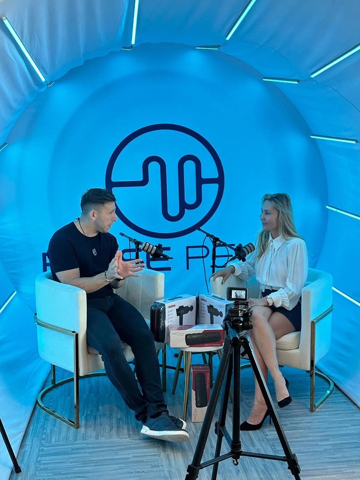 Two people are seated in a blue-lit studio with a large logo behind them, engaging in a conversation with microphones. There are boxes of audio equipment on a small table between them and a camera set up in front.