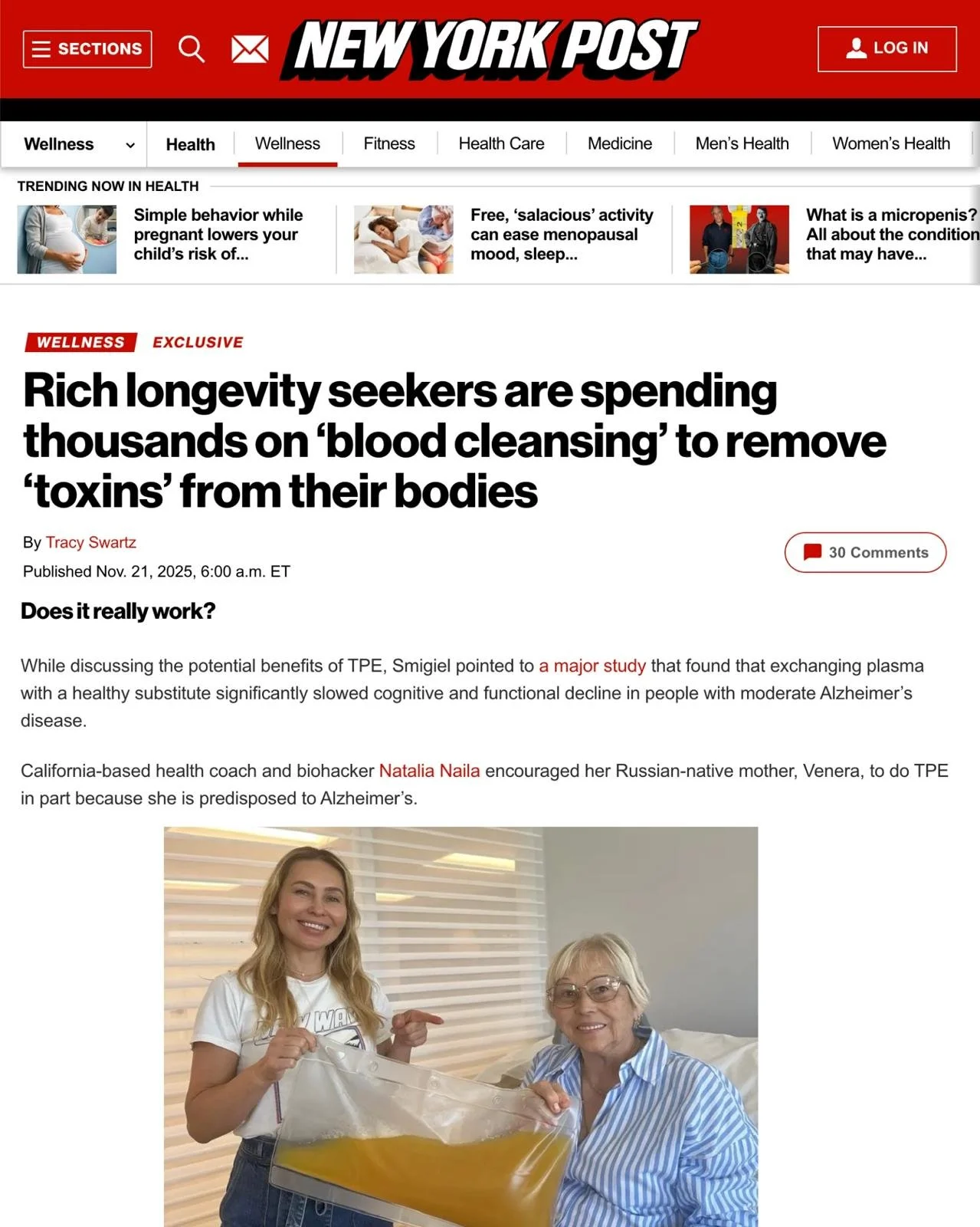 A smiling woman holding a large plastic bag filled with yellow liquid or gel in front of an elderly woman with glasses, who is sitting on a bed in a well-lit room with a window and blinds.