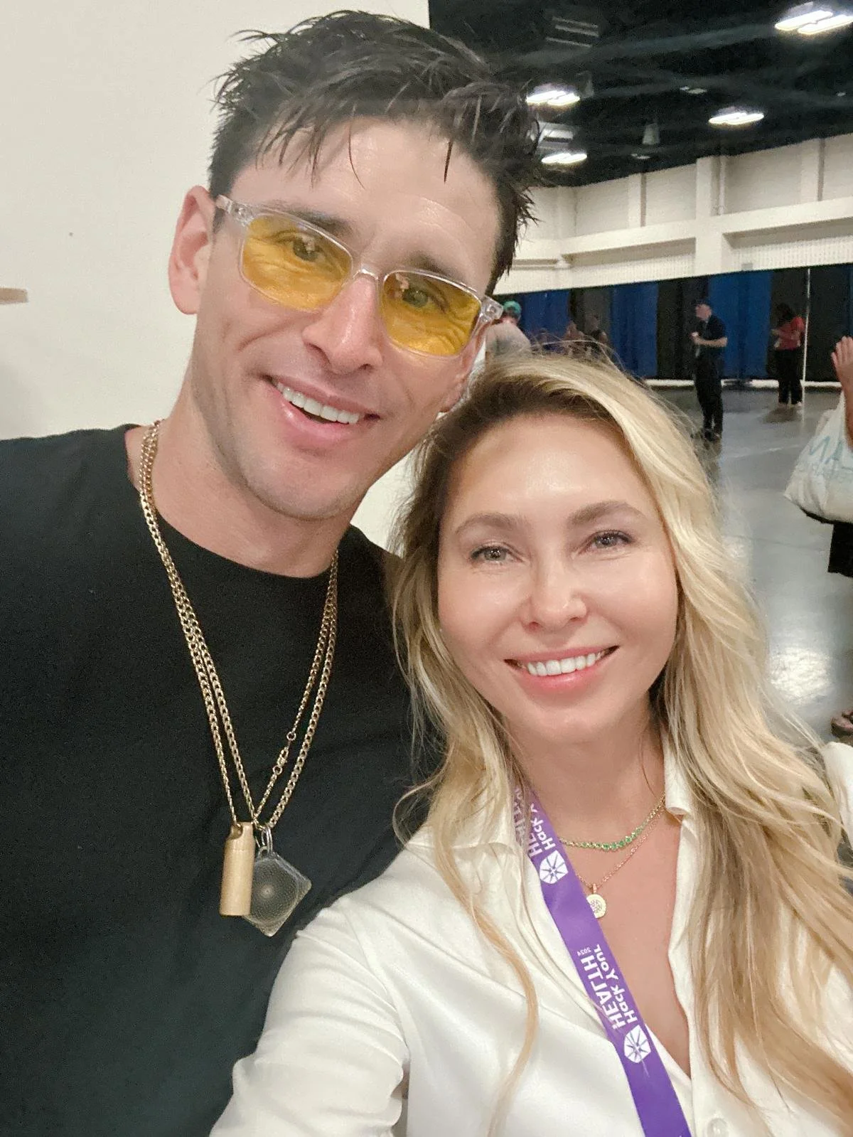 A man and woman taking a selfie at an indoor event. The man is wearing yellow-tinted glasses, a black shirt, and gold necklaces. The woman has blonde hair, is smiling, and is wearing a white shirt with a purple lanyard around her neck.