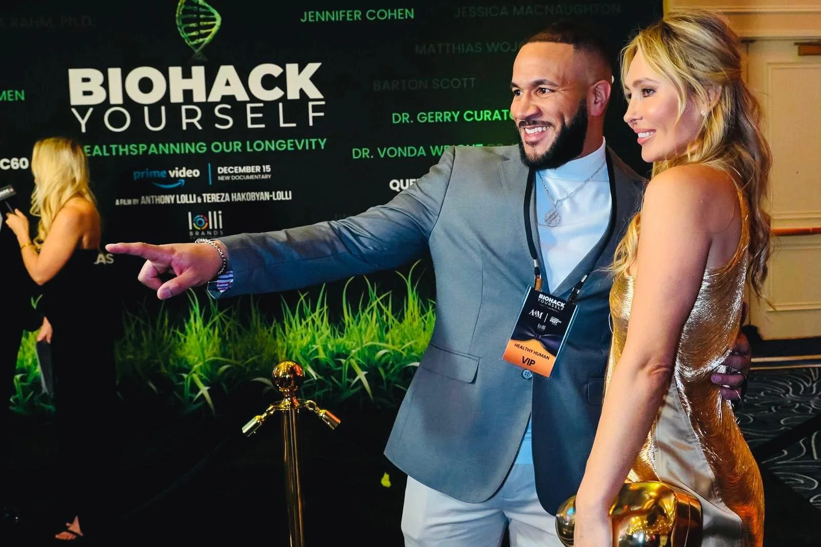 A man and woman taking a selfie at a conference or event, with a large poster behind them promoting a documentary called 'Biohack Yourself.' The man is pointing at the poster while the woman smiles, holding a golden clutch purse. The man is wearing a