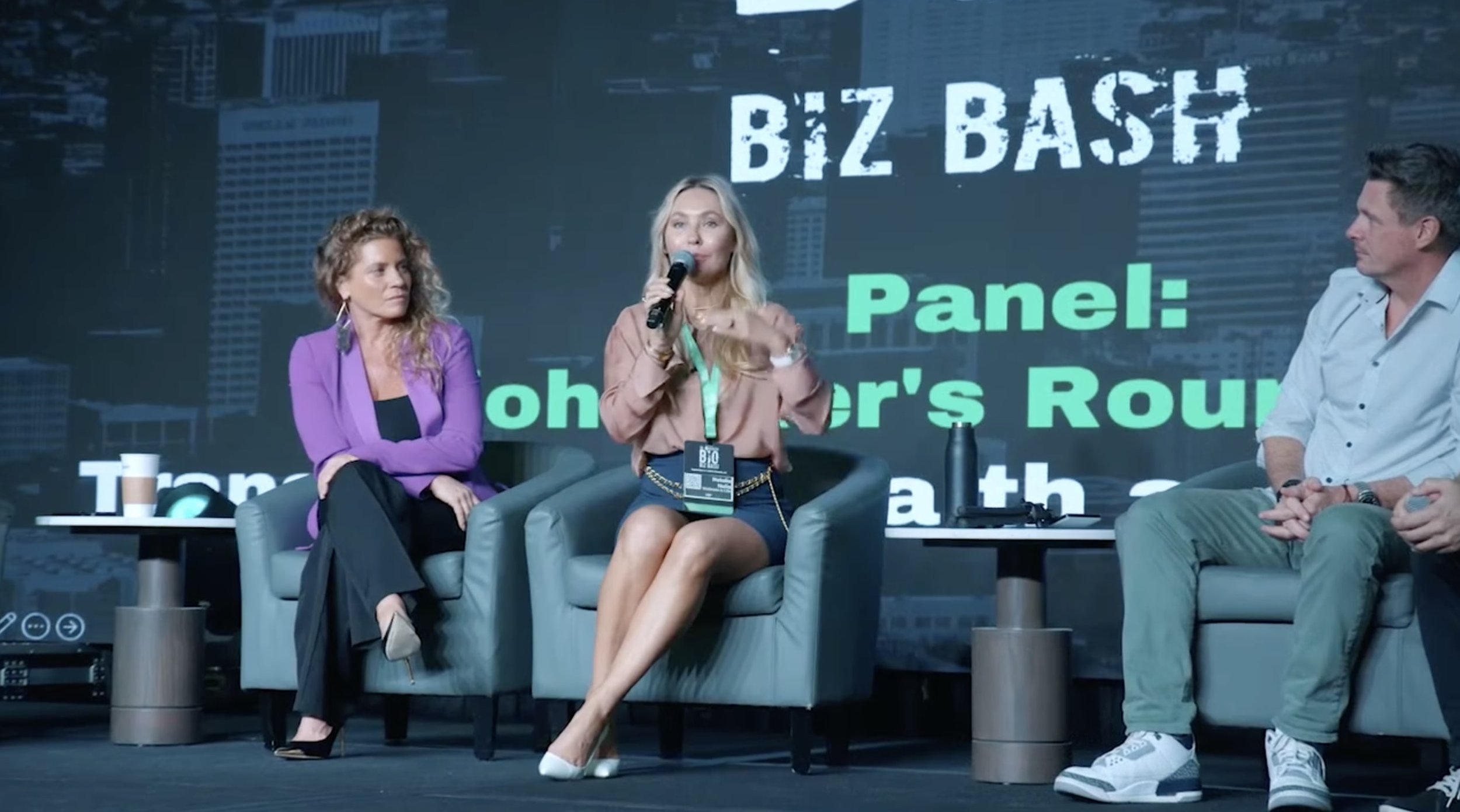 Three people sitting on stage at Biz Bash event, with a large screen behind them displaying 'Biz Bash Panel:...er’s Round'.