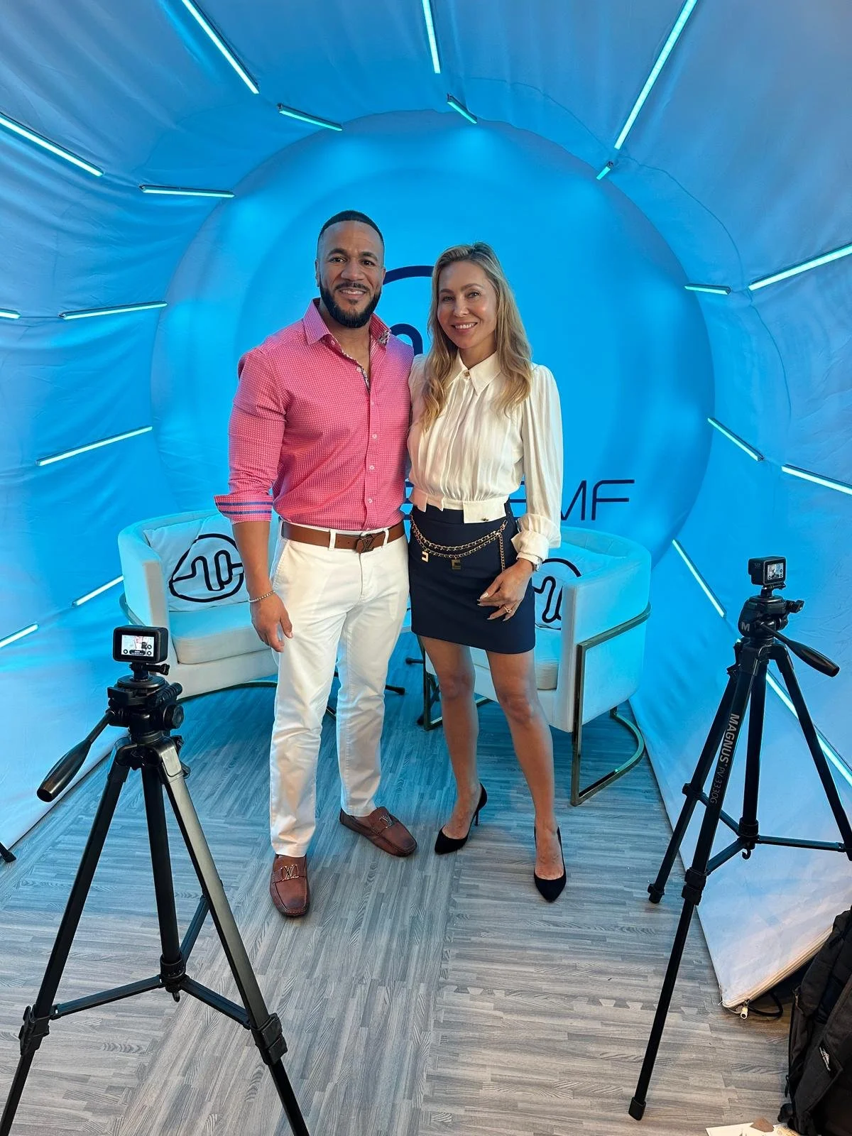 A man and woman standing together in a photo booth with a blue background, two chairs, and camera equipment. The man is wearing a pink checkered shirt and white pants, and the woman is wearing a cream blouse and black skirt with high heels.