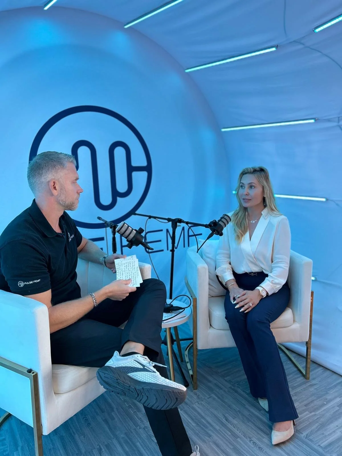 A man and a woman are sitting in an interview setup with microphones. The man is holding a note, and they are seated on white chairs inside a tent with blue lighting. There is a logo behind them.