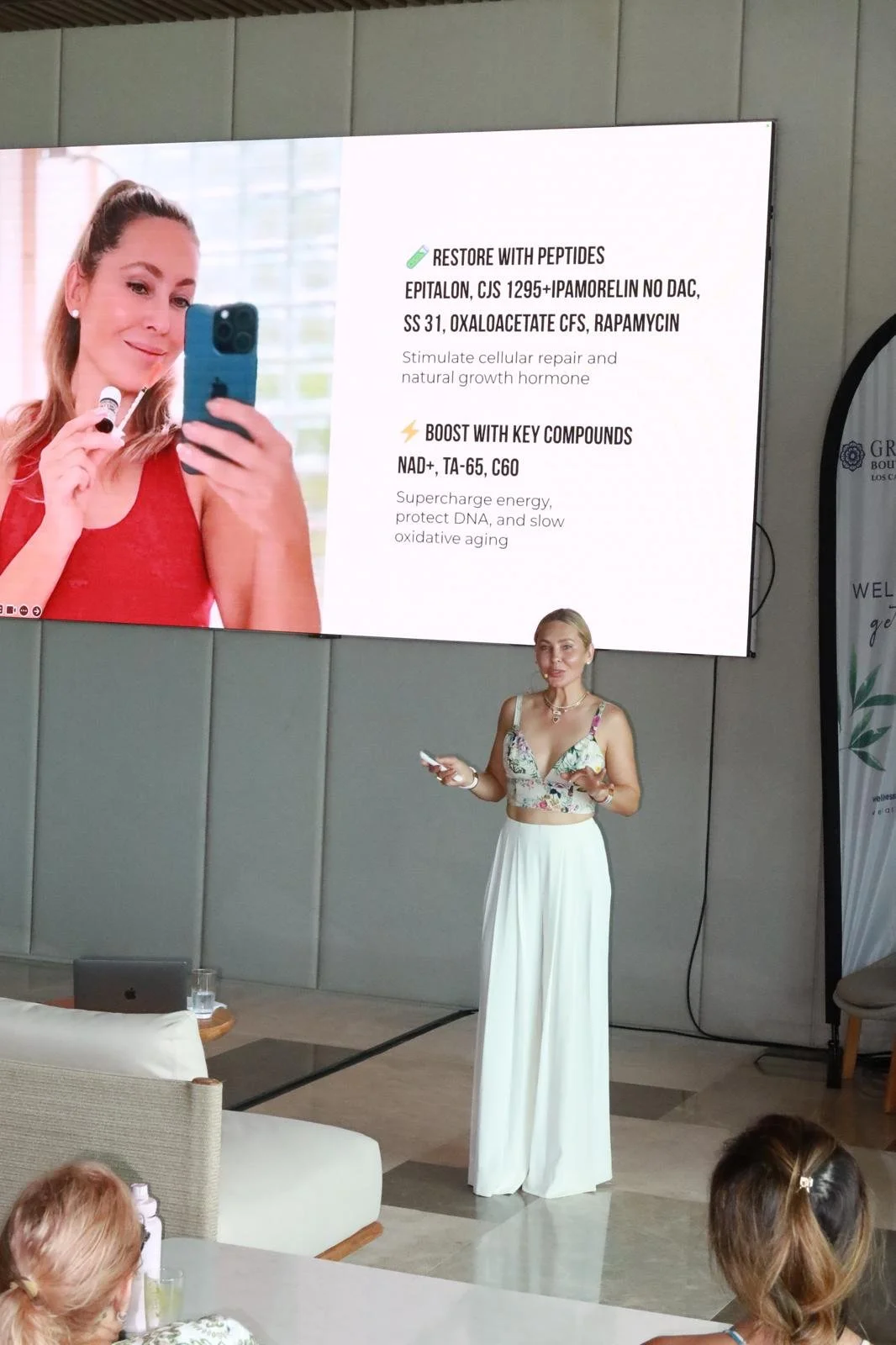 A woman in a floral crop top and wide-leg white pants giving a presentation in front of a large screen displaying skincare product information. The screen shows a woman holding a skincare product and lists ingredients and benefits.