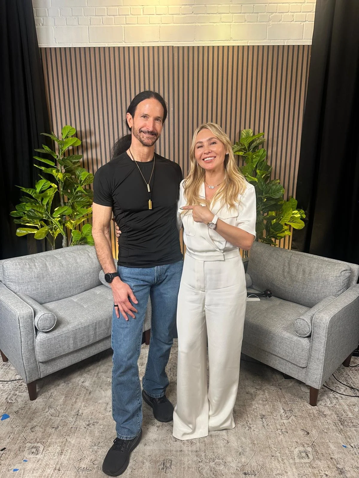 A man with long dark hair, a beard, wearing a black t-shirt and jeans, standing next to a blonde woman in white outfit; both smiling and posing in front of a patterned wall with two potted plants and a gray couch.