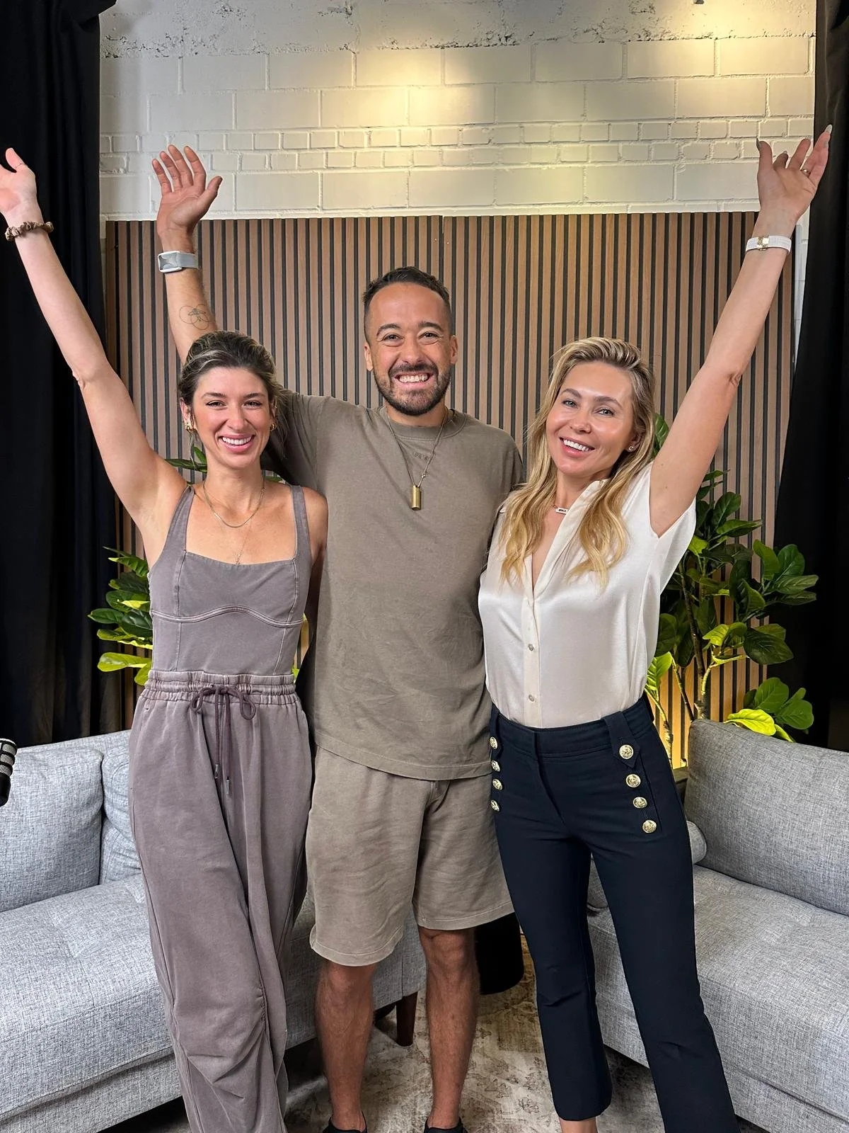 Three people standing together indoors, smiling, with arms raised and hands open, in front of a wooden wall with green plants on either side.