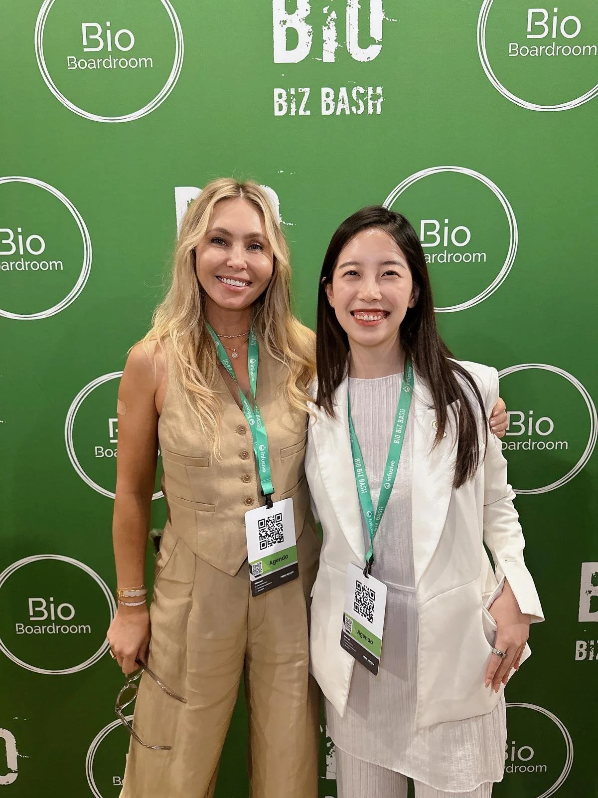 Two women smiling at a BIO Boardroom event, standing in front of a green backdrop with 'Bio Boardroom' and 'BIZ BASH' logos, both wearing conference badges.
