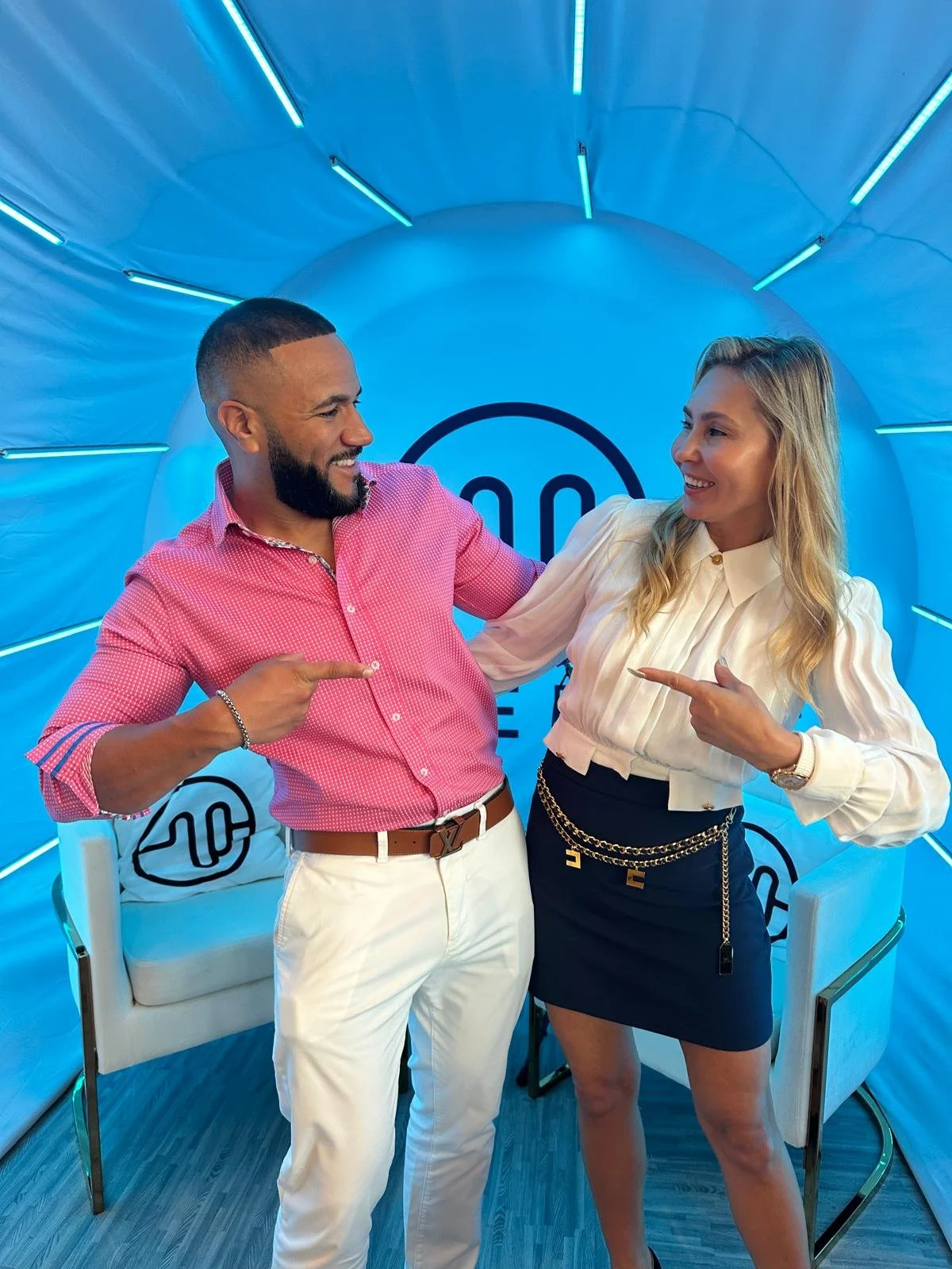 A man and woman pointing at each other smiling in a colorful, modern studio with a blue backdrop and seating area.