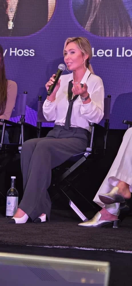 Woman with blonde hair in a white blazer and gray wide-leg pants sitting on a stage, holding a microphone and speaking, with a large screen behind her showing text and part of another person's image.