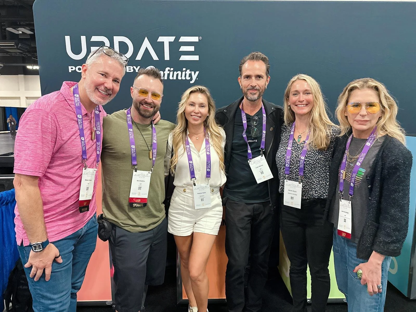 Six people standing together at a conference, smiling. They are wearing conference badges and lanyards. Behind them is a large sign with the words 'UPDATE Powered by Microsoft and Xfinity'.