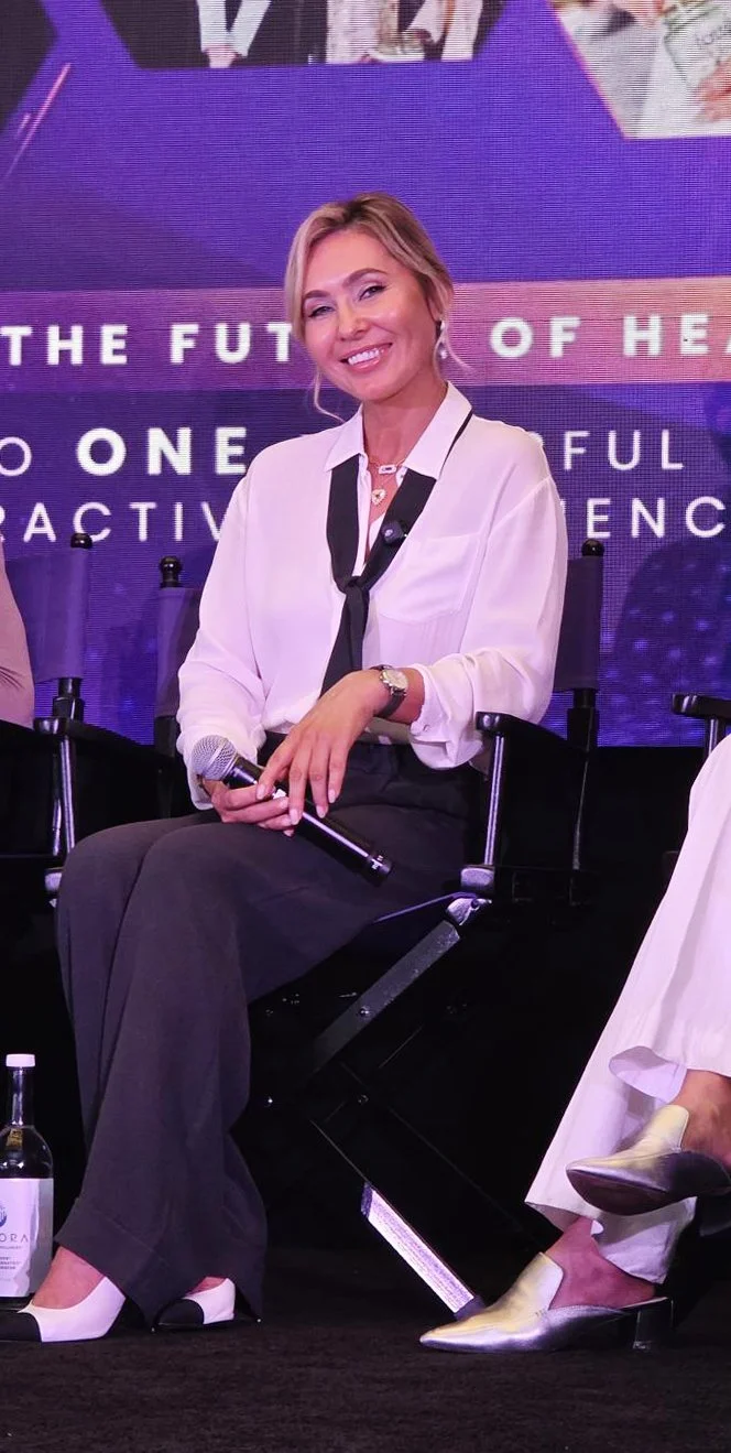A woman with blonde hair in a white blouse and black pants, sitting on a black chair, holding a microphone, smiling, at a panel or conference with a purple background.