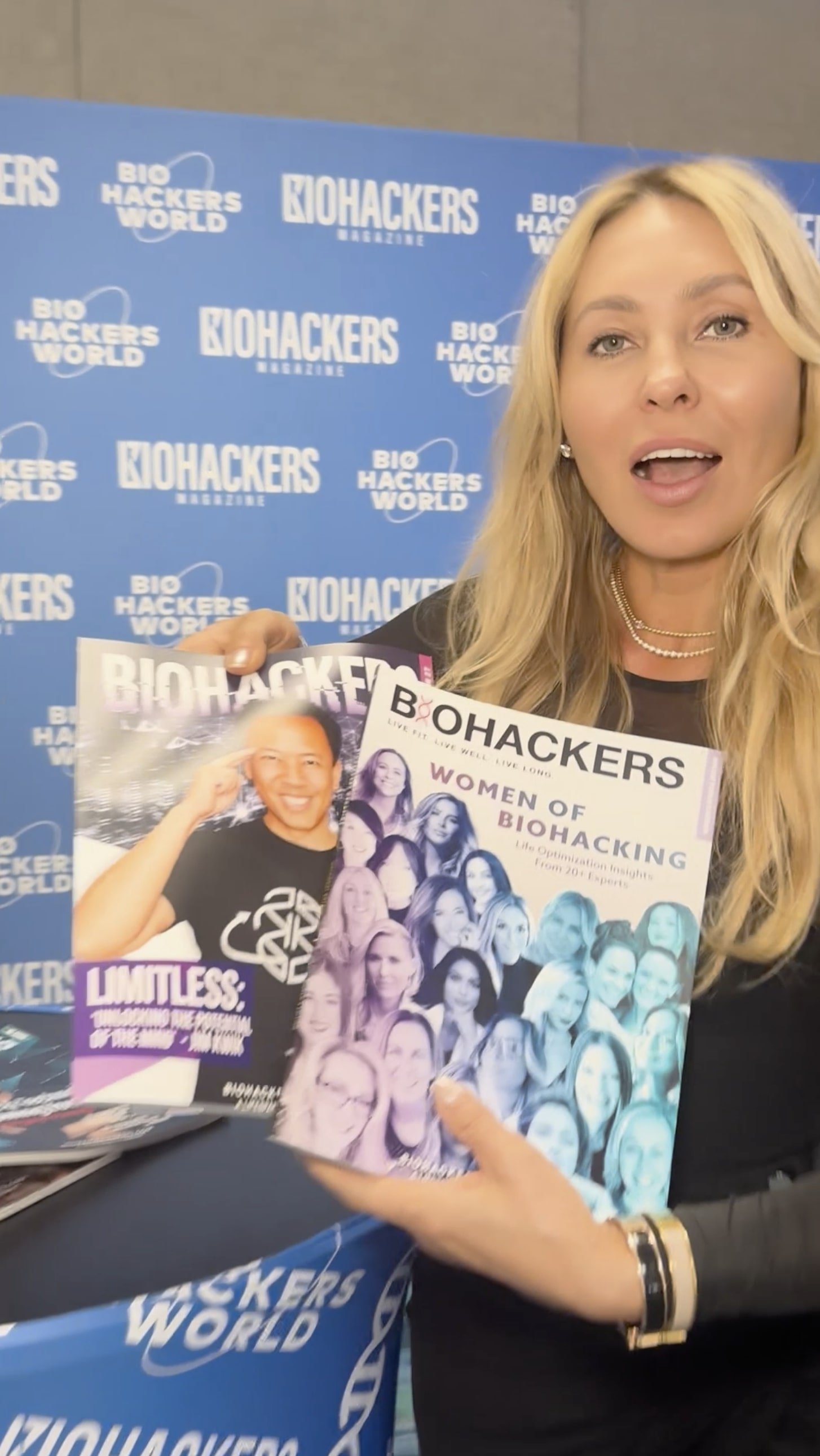 A woman with long blonde hair holding biohacking magazines at a Bio Hackers event, with a blue backdrop covered in Bio Hackers branding.