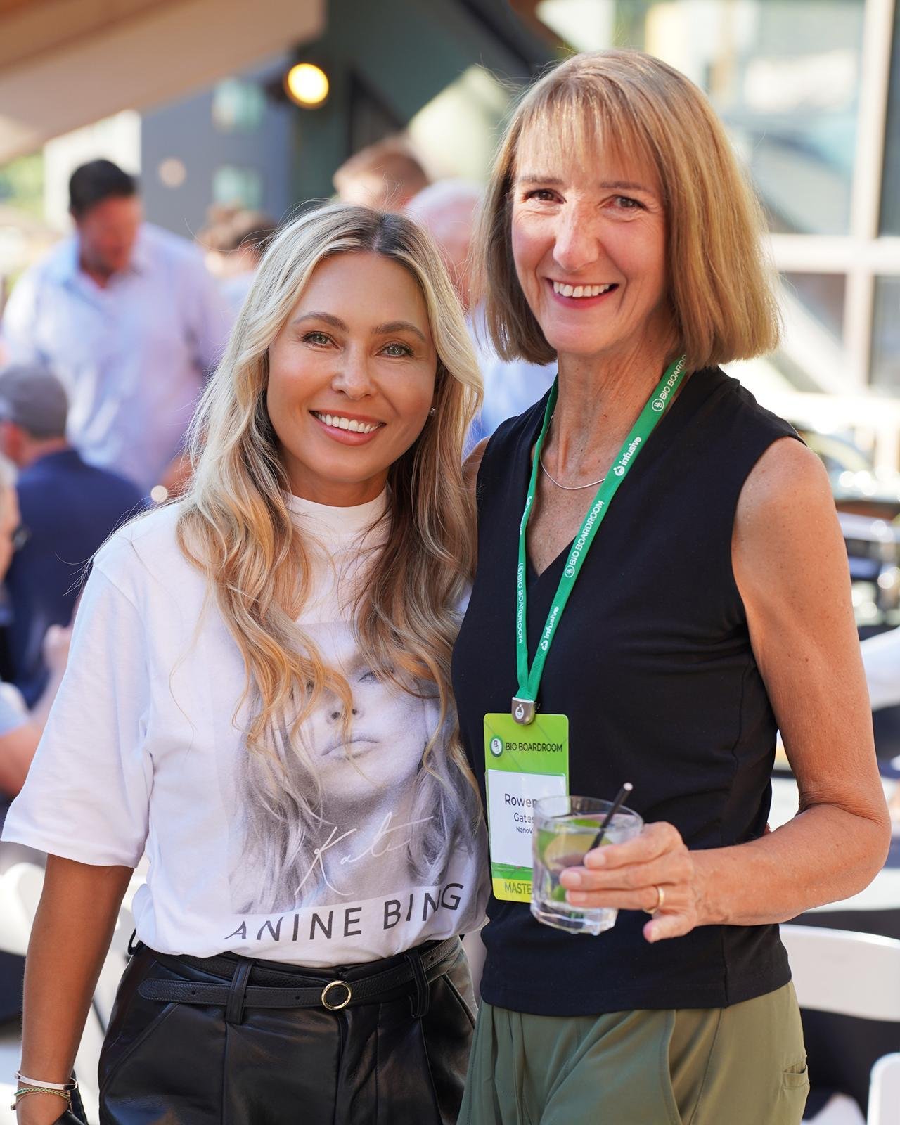 Two women smiling at an outdoor event, one woman wearing a white graphic t-shirt and the other in black sleeveless top with a green lanyard, holding a drink.