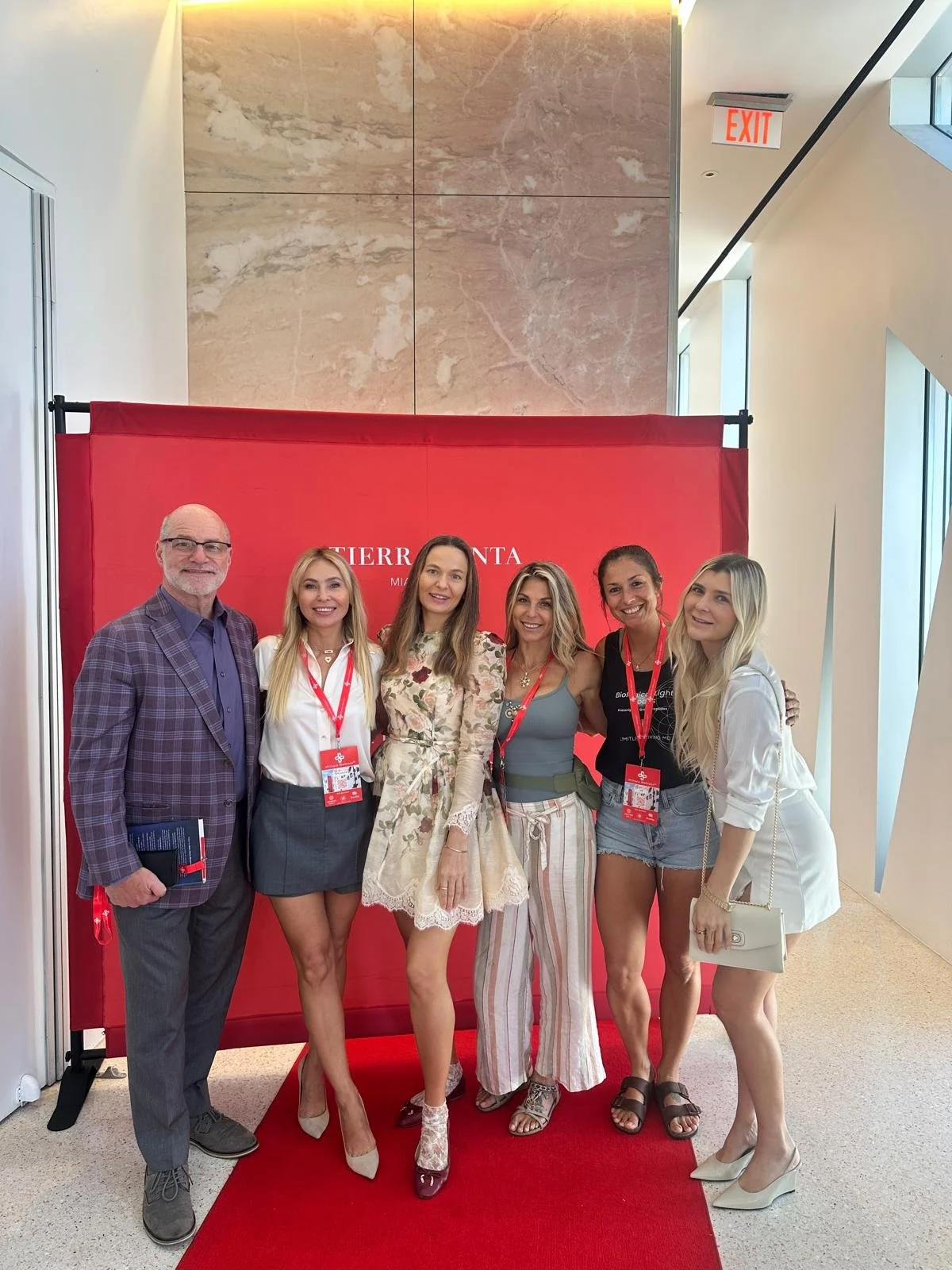 Six people standing in front of a red backdrop with white text, smiling at the camera in a modern indoor setting with marble and glass walls.