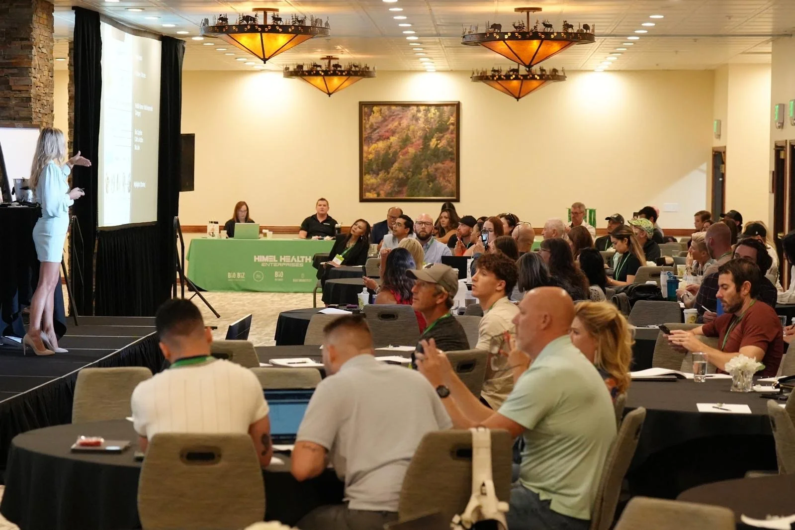 A woman in a white outfit is giving a presentation to a large audience in a conference room with round tables, some with laptops and notebooks, and a green table with a banner that reads 'HIMEL HEALTH'. The room has warm lighting and a large framed p