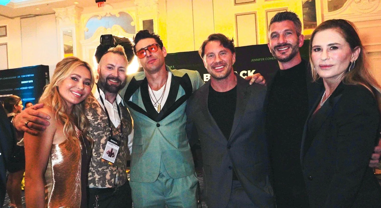 Group of six actors and actresses posing together at a promotional event, smiling, with a backdrop and room decor indicating a formal or entertainment industry event.