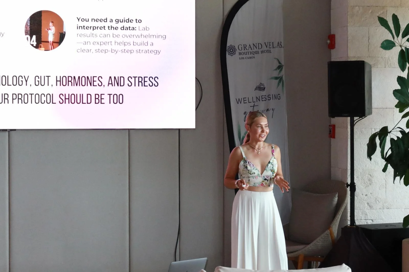 A woman giving a presentation in front of a large screen at Grand Velas Boutique Hotel in Los Cabos. The screen displays text about interpreting data, hormones, and stress. A banner with the word 'WELLNESS' and some greenery decor are also in the bac