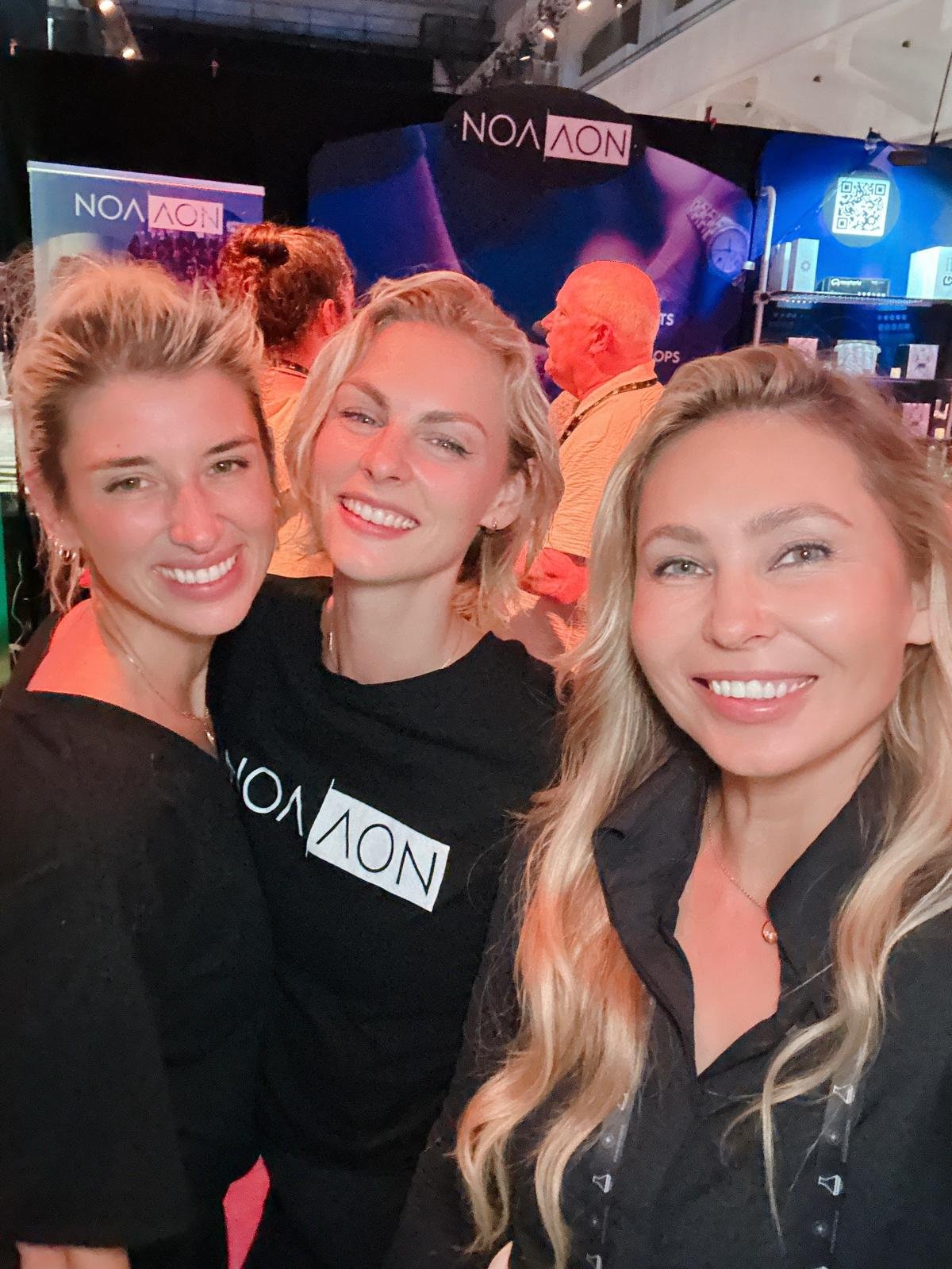 Three women smiling at a scene with a Navi logo in the background, likely at an event or conference.