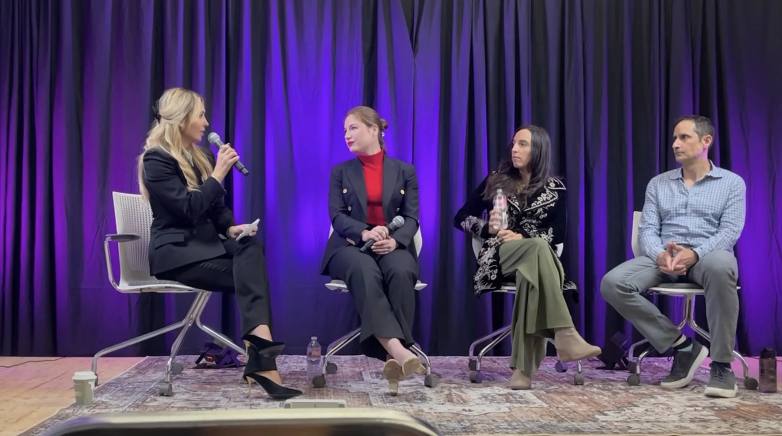 Four people sitting on chairs on a stage in front of a purple curtain, engaged in a panel discussion. The woman on the far left is speaking into a microphone, wearing a black suit and high heels. The second woman from the left has red hair, is dresse