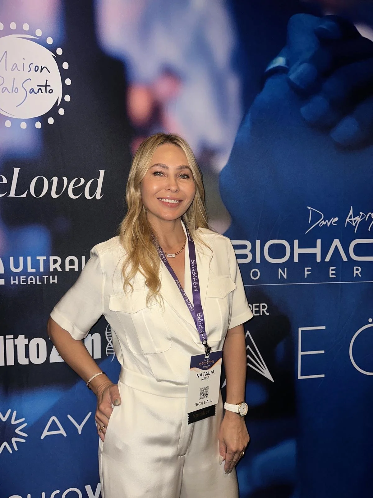 A woman with blonde hair, wearing a white outfit and a white watch, standing in front of a blue and black backdrop with event logos, smiling at the camera.