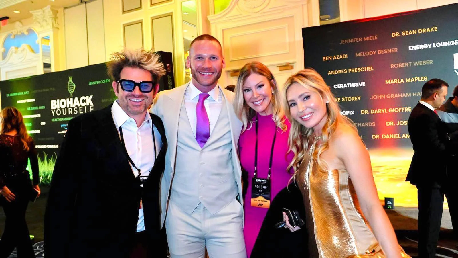 Four people smiling at a conference with a Biohack Yourself backdrop. The group includes one man with glasses and wild hair, a man in a white suit with a purple tie, a woman with blonde hair in a pink outfit, and a woman in a shiny gold dress.