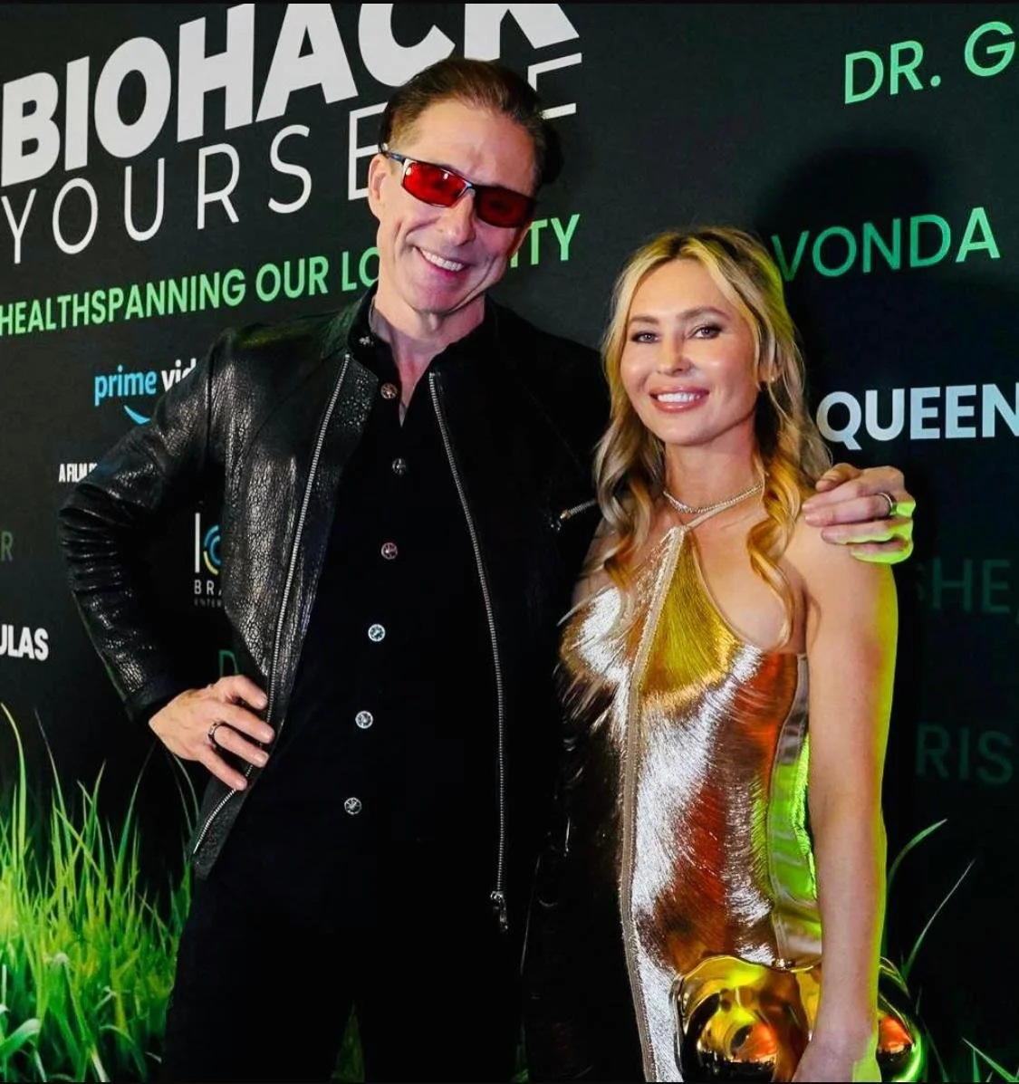 A man and a woman posing together at an event, smiling. The man is wearing red sunglasses, a black leather jacket, and a black shirt. The woman is wearing a shiny, metallic gold dress and a necklace. The background has text related to biohacking and 