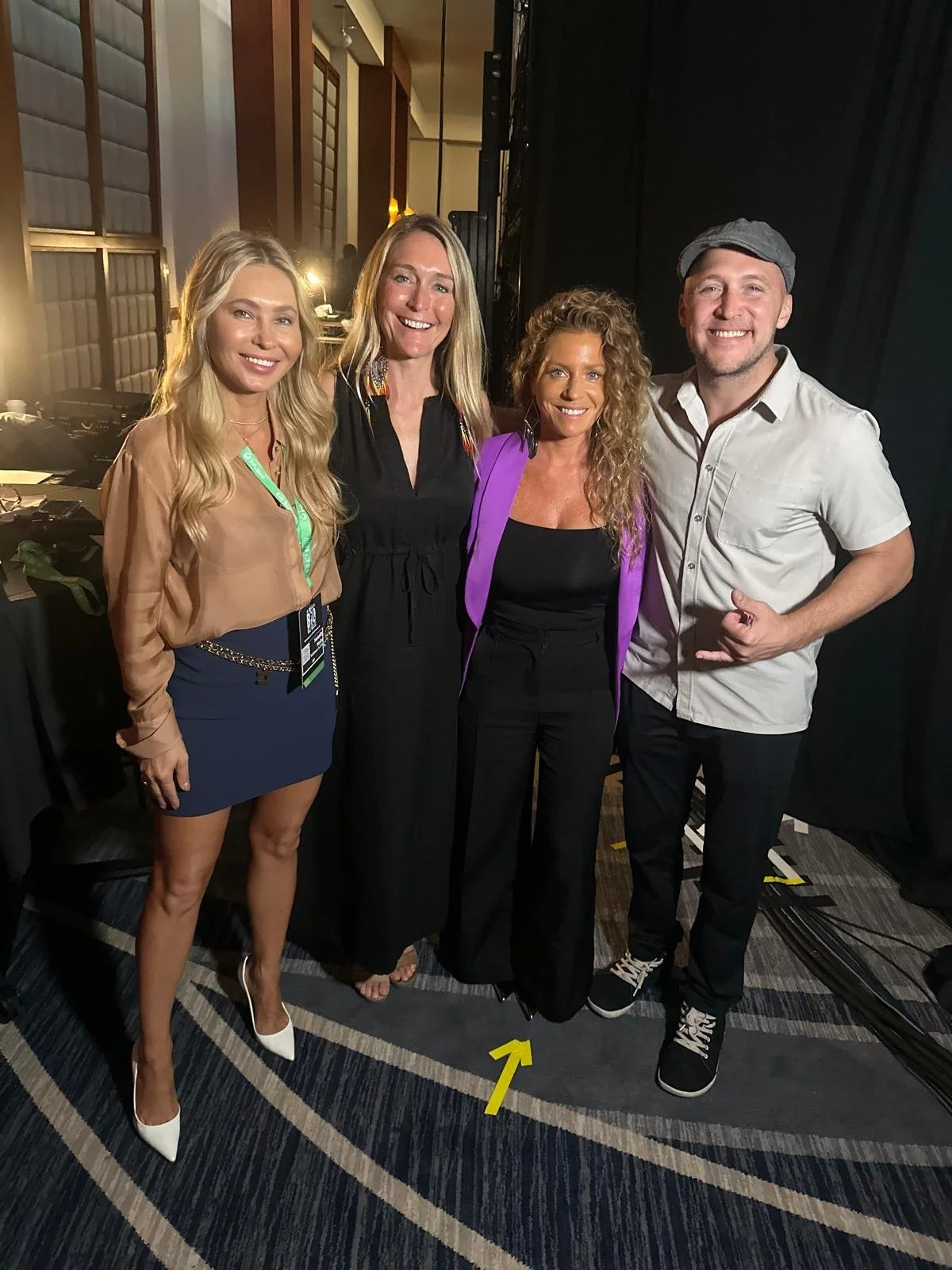 Four people standing together indoors, smiling at the camera. Three women and one man, dressed in stylish attire, with a black curtain in the background and stage lighting.