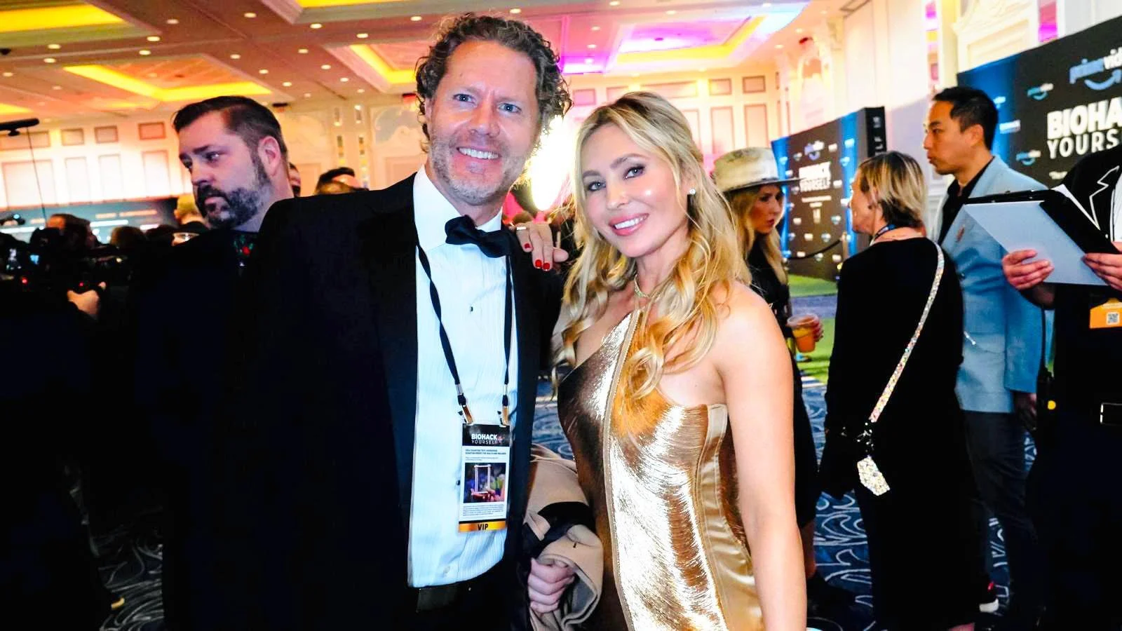 A man in a tuxedo and a woman in a gold dress smiling at the camera at an event with a colorful, decorated background.