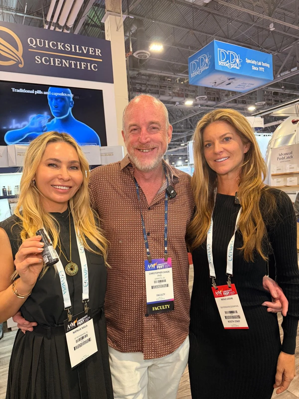 Three people smiling and posing for a photo at a scientific conference. The man in the middle is wearing a brown patterned shirt with a conference badge that says "Faculty". The woman on the left is holding a small bottle and has a badge that reads "