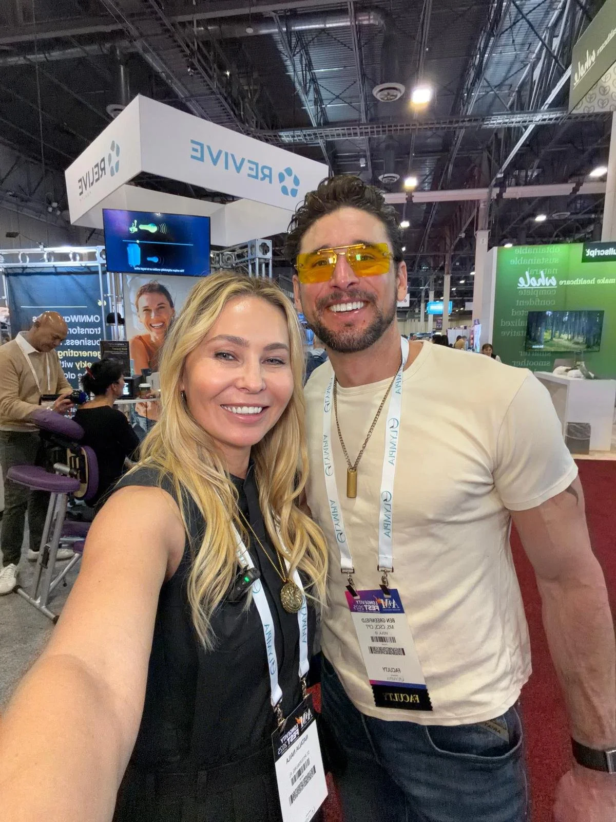 Two smiling adults taking a selfie at a conference or trade show. The woman has long blonde hair and is wearing a black sleeveless top, while the man has short dark hair, facial hair, yellow-tinted sunglasses, a white t-shirt, and a badge around his 