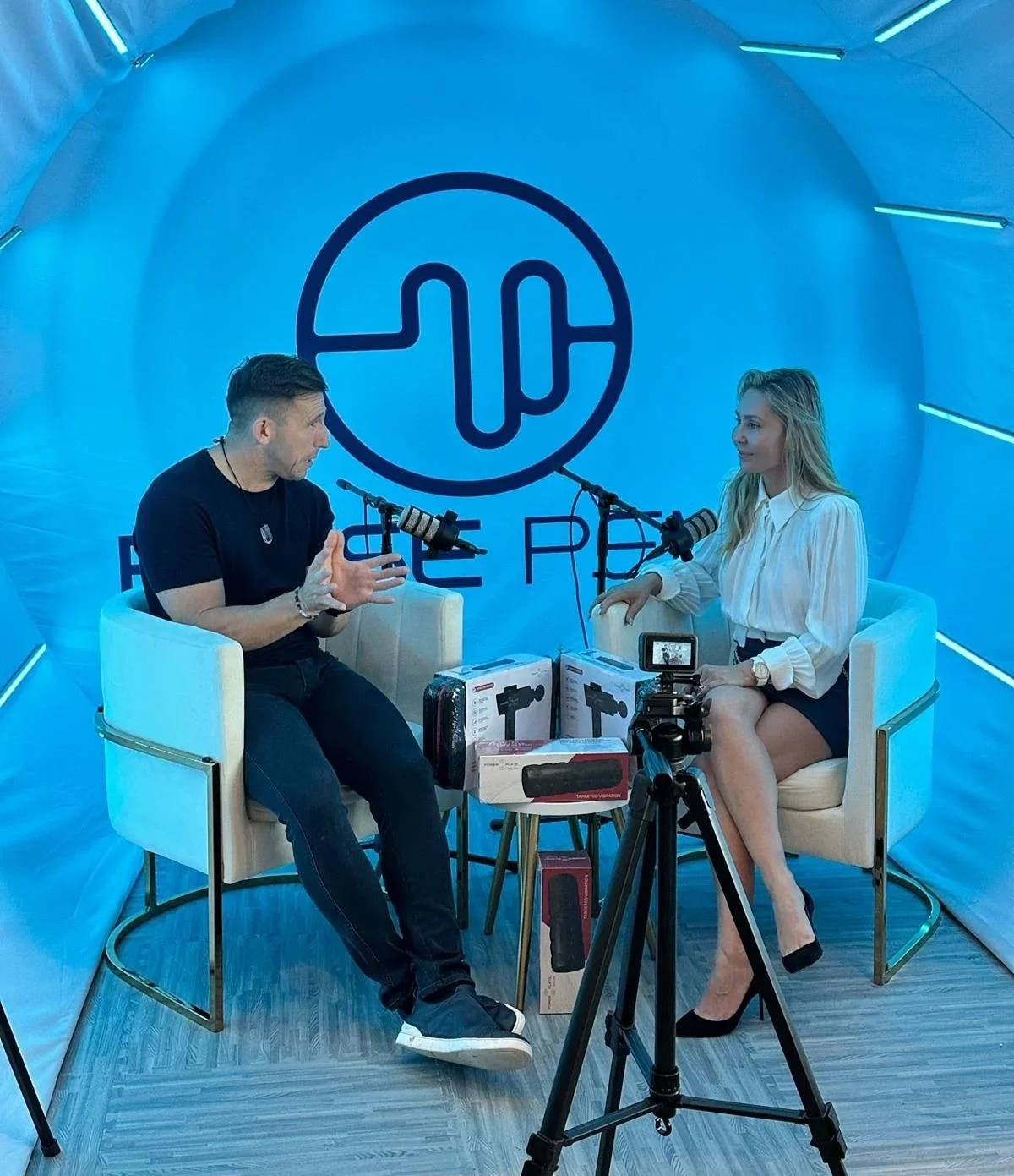 Two people in a blue-tinted recording studio with a large logo behind them, microphones, camera, and boxed microphones on a small table between them, engaged in conversation.